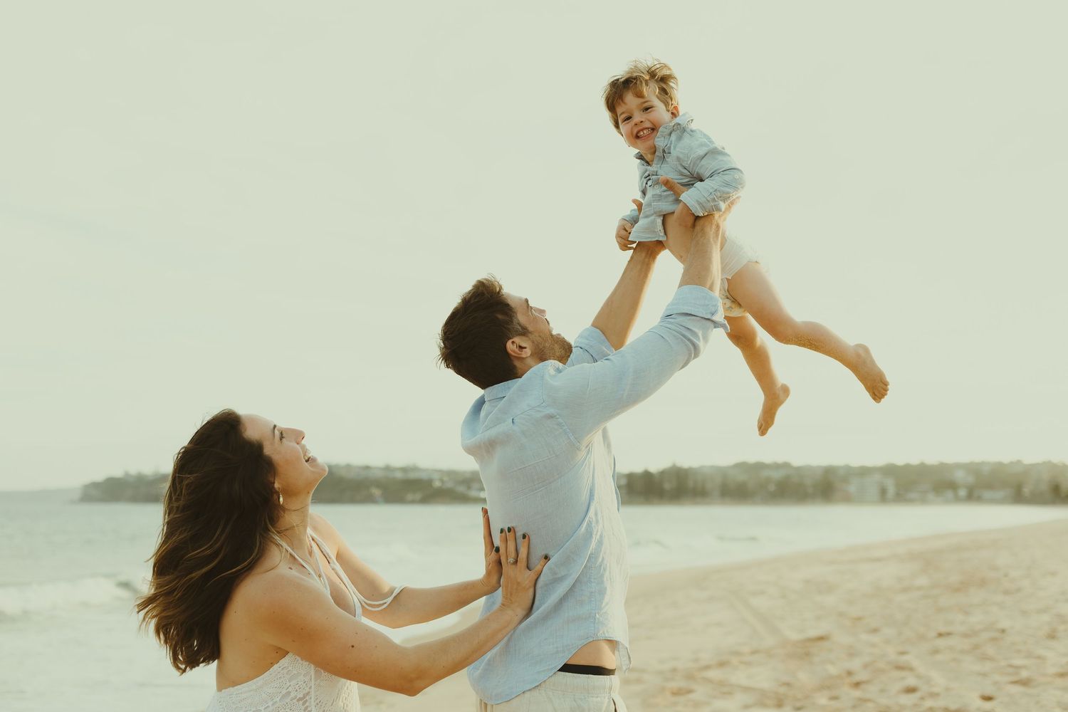 A family plays and lifts their child in the air while enjoying time at the beach.
