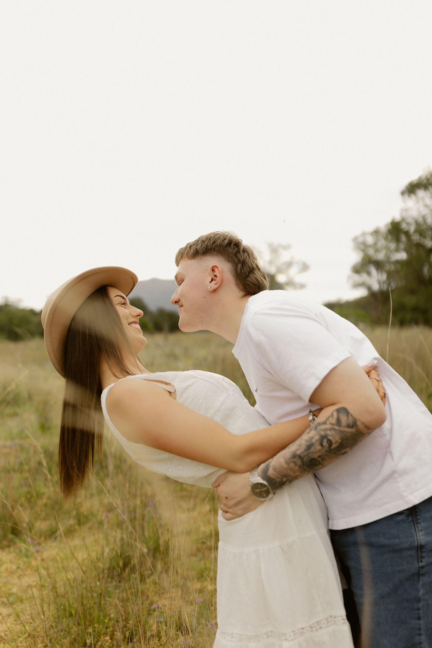 A romantic couple shares playful kisses in a rustic field with mountains in the background.