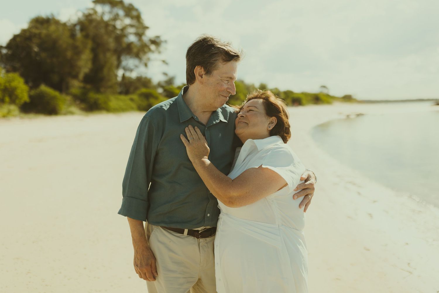 A tender moment between a couple embracing on a scenic beach with trees in the background.