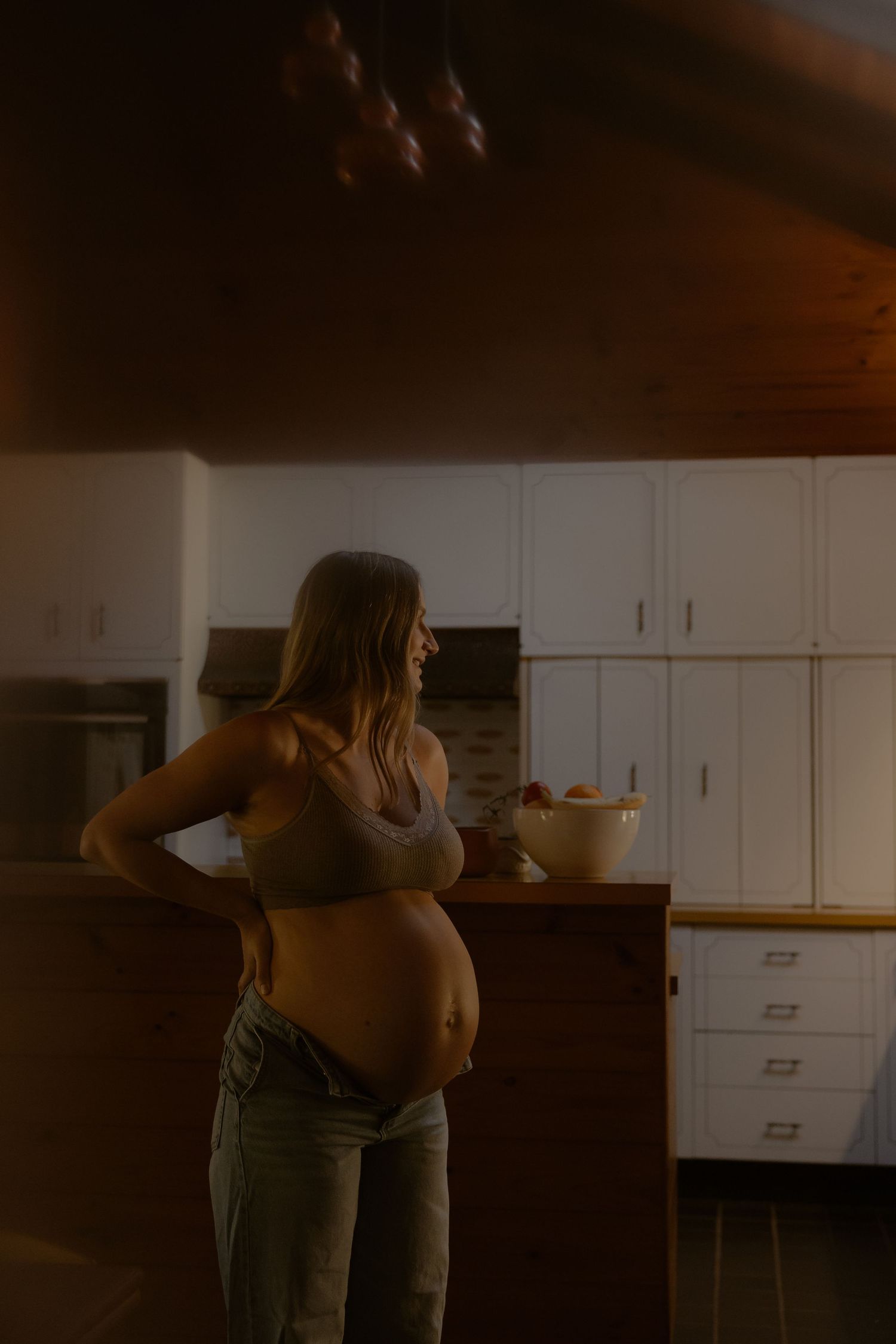 Pregnant woman in bra and jeans standing in a warmly lit kitchen cradling her baby bump.