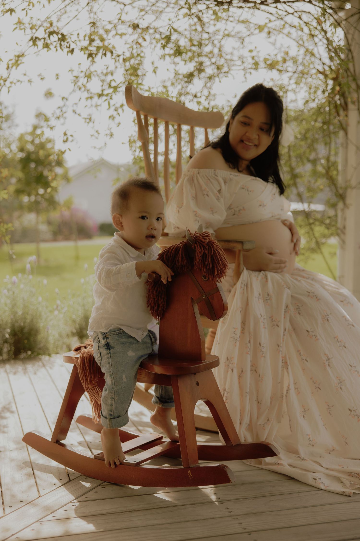Pregnant woman in floral dress sits in rocking chair beside toddler on wooden rocking horse on sunlit porch.