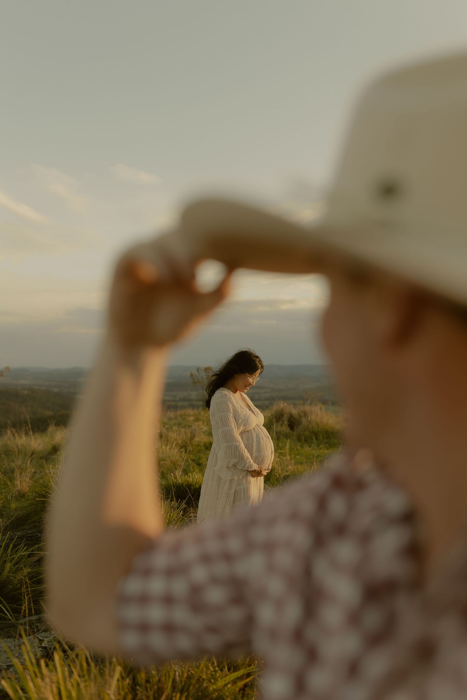 Person in checkered shirt and hat frames pregnant woman in white dress on sunlit hill.