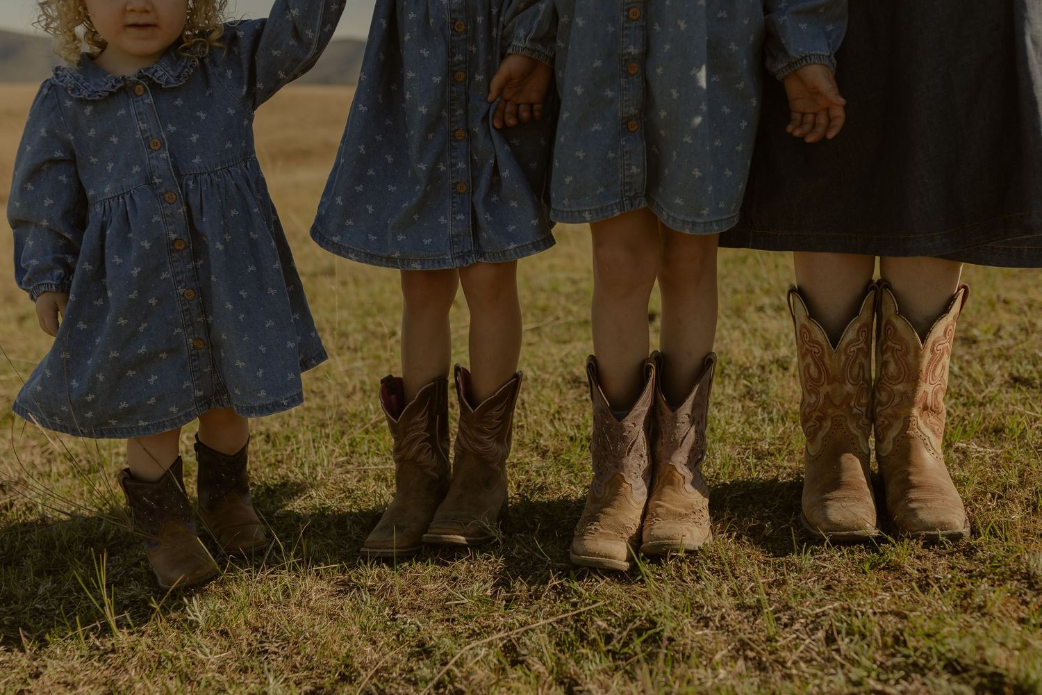 Three young girls in blue floral dresses and cowboy boots standing on grass in warm sunlight.