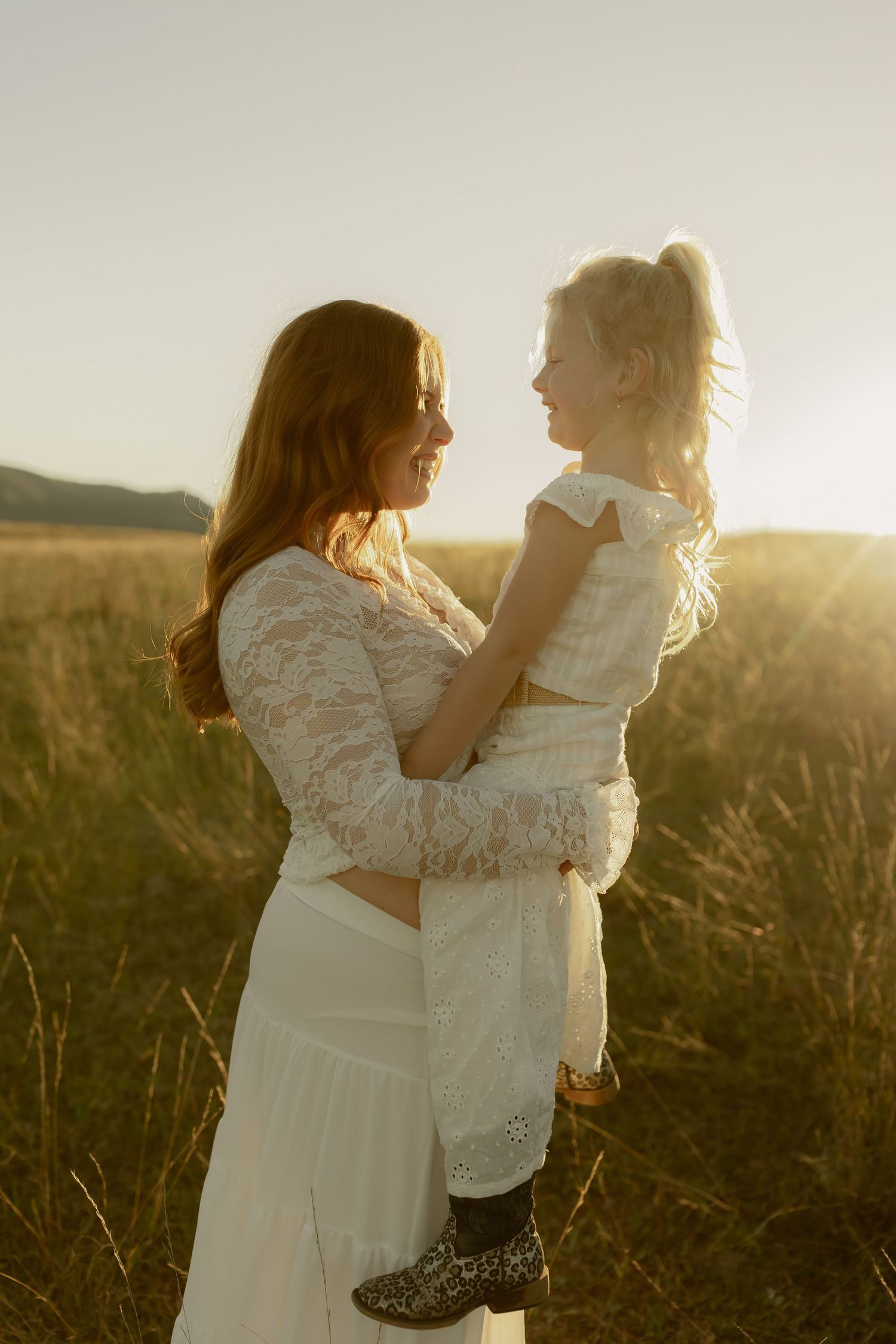 A mother in white lace holds her young blonde daughter in a sunlit golden field at sunset.