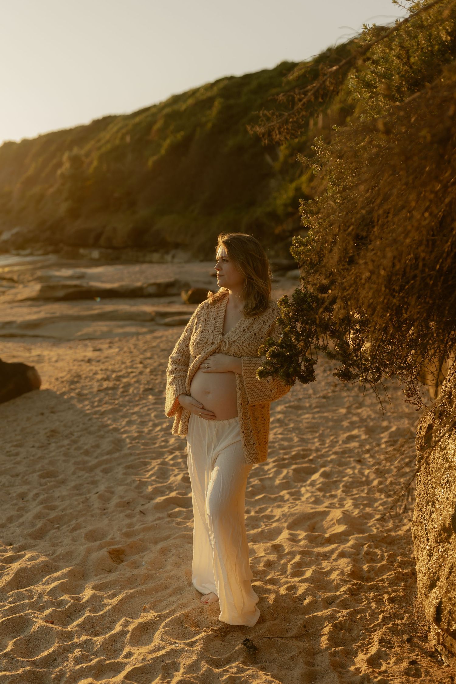 Pregnant woman in white dress and cardigan holding belly at golden hour beach sunset.