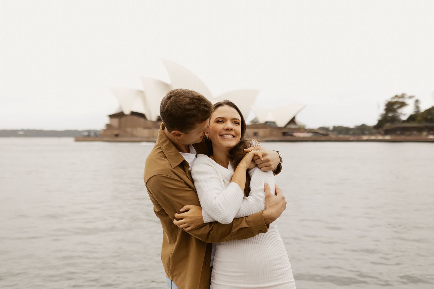 A couple embraces by the water with the Sydney Opera House visible in the background.
