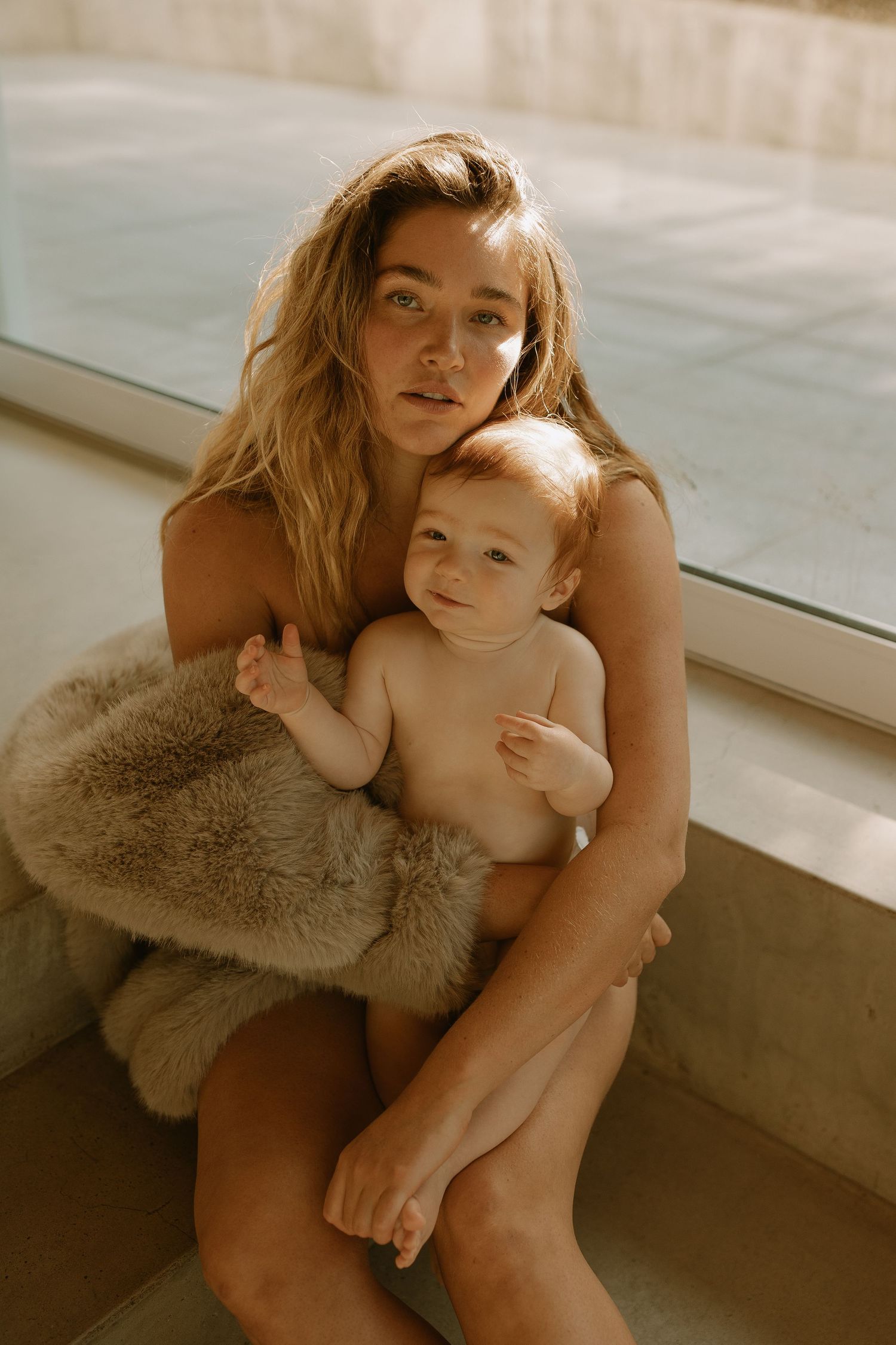 Intimate portrait of someone holding a baby against a window with warm sunlight and a cozy fur blanket.