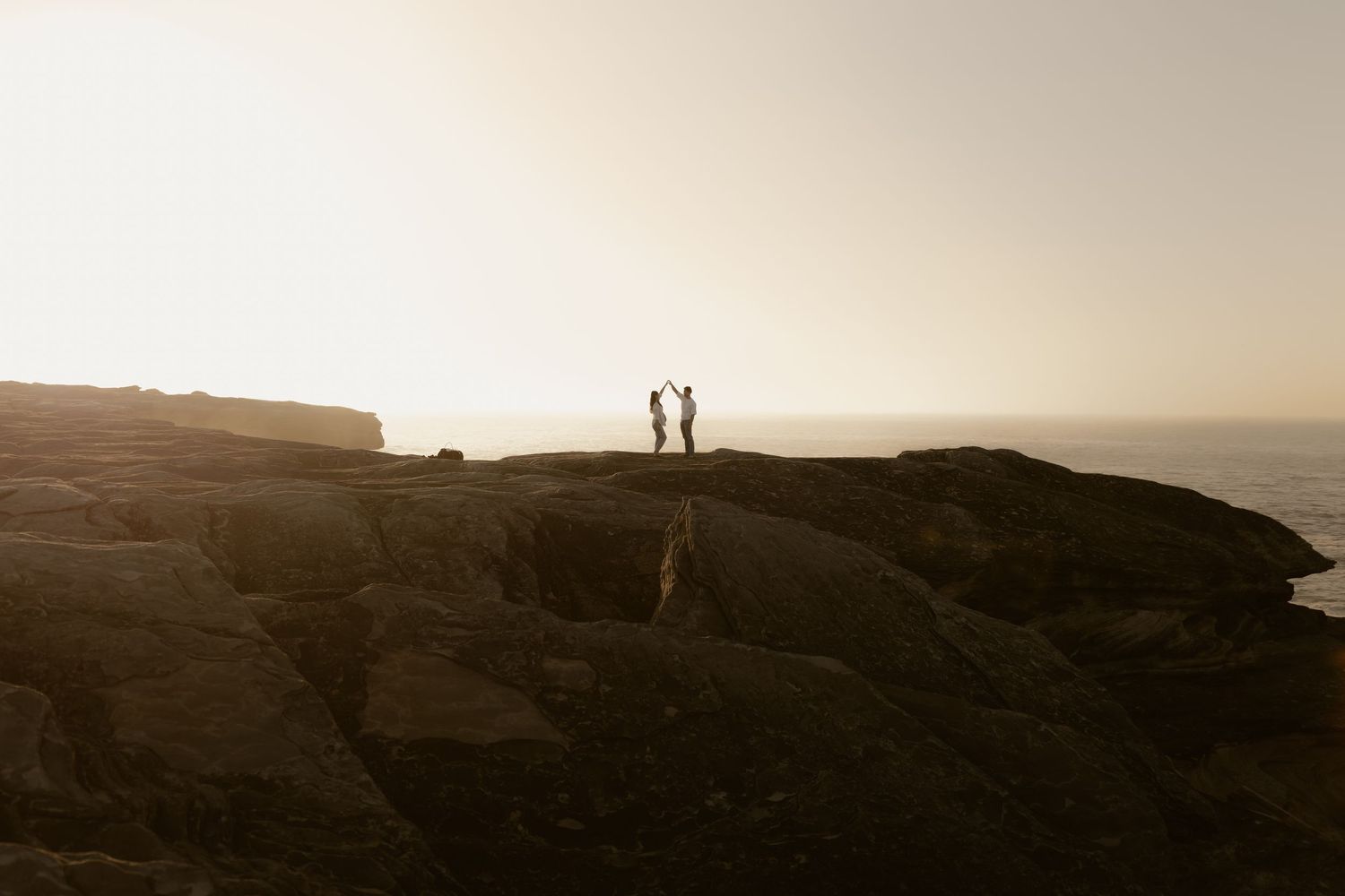 Silhouettes of people standing on coastal cliffs against a misty sunset sky.