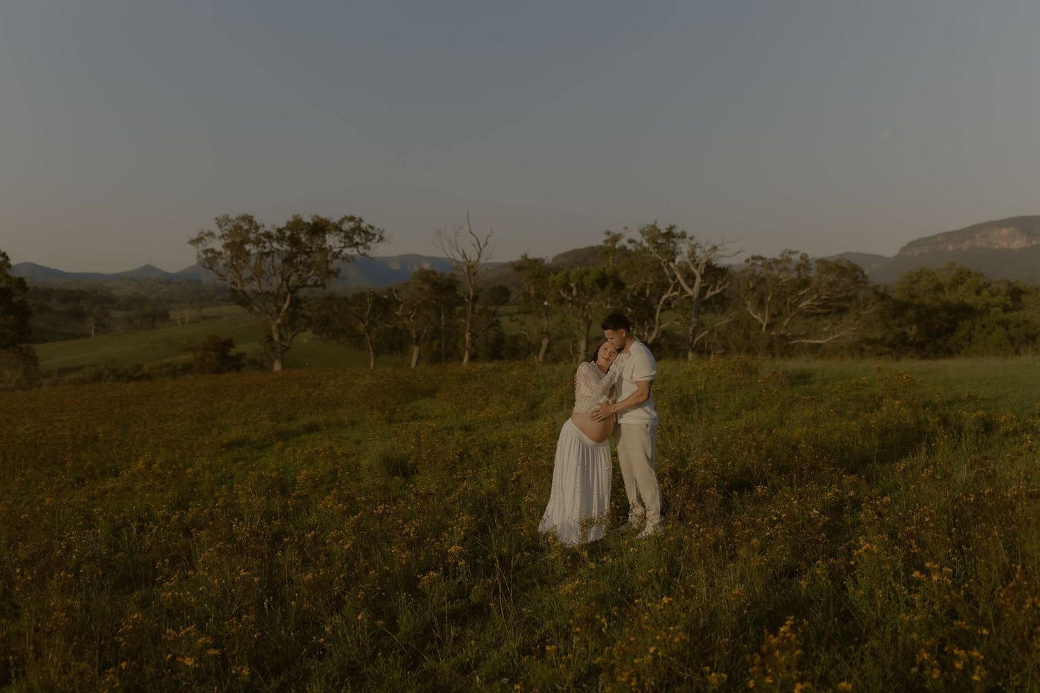 Two people stand close together in a vast field of flowers with rolling hills in the distance.