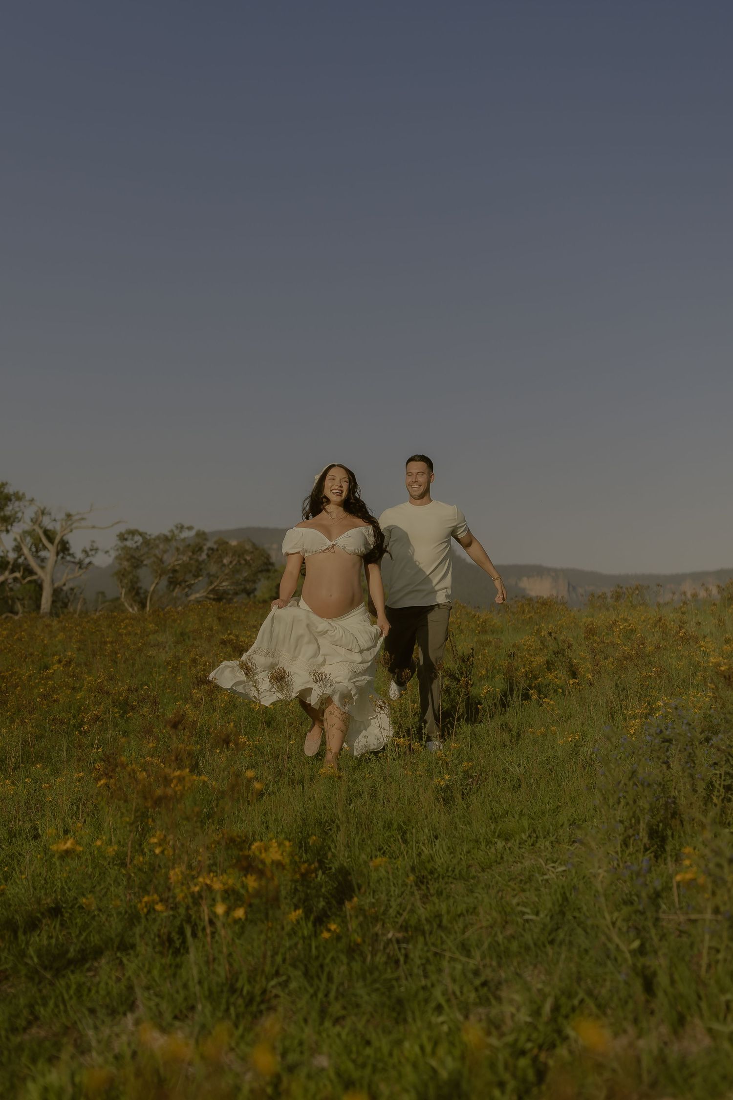 A couple runs together through a sunlit meadow with mountains in the background during golden hour.