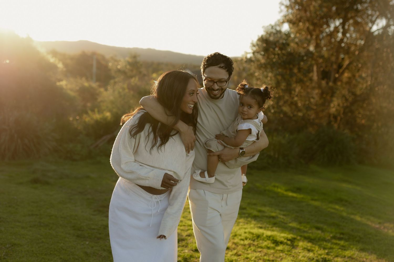 Family of three enjoying a golden sunset outdoors, parents smiling at toddler in white outfits on green grass.