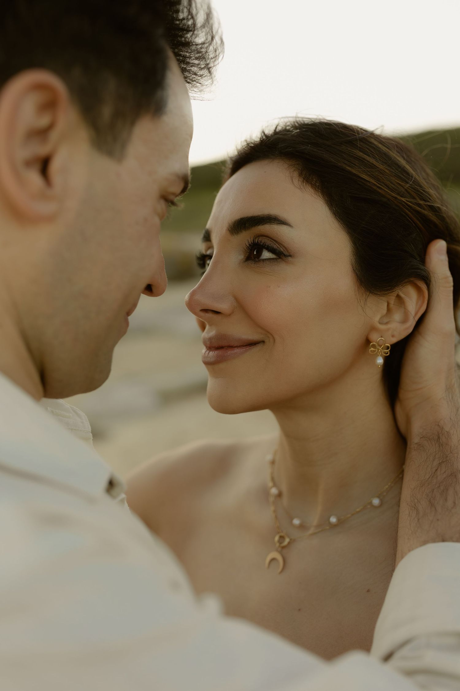 Close-up of couple almost kissing, woman wearing gold crescent necklace and earrings at golden hour.