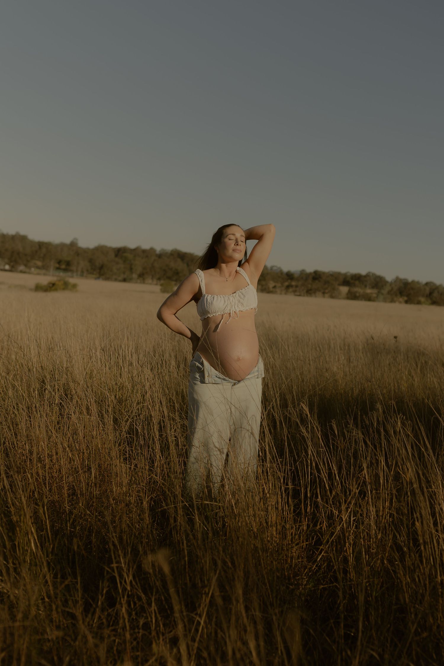 Pregnant woman standing in golden field wearing white crop top and pants, posing outdoors at dusk.