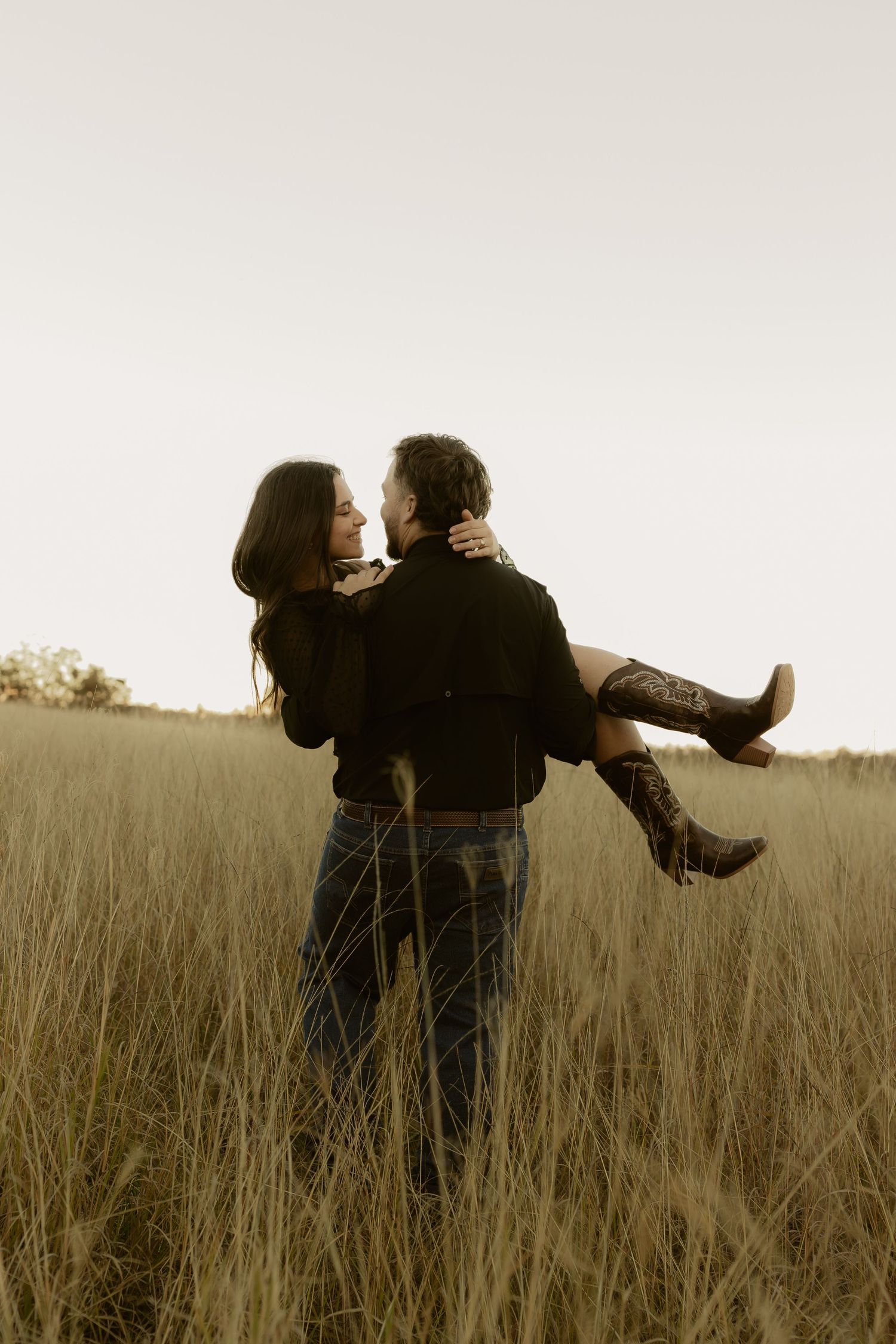 Man carrying woman in his arms through a tall golden grass field at sunset.