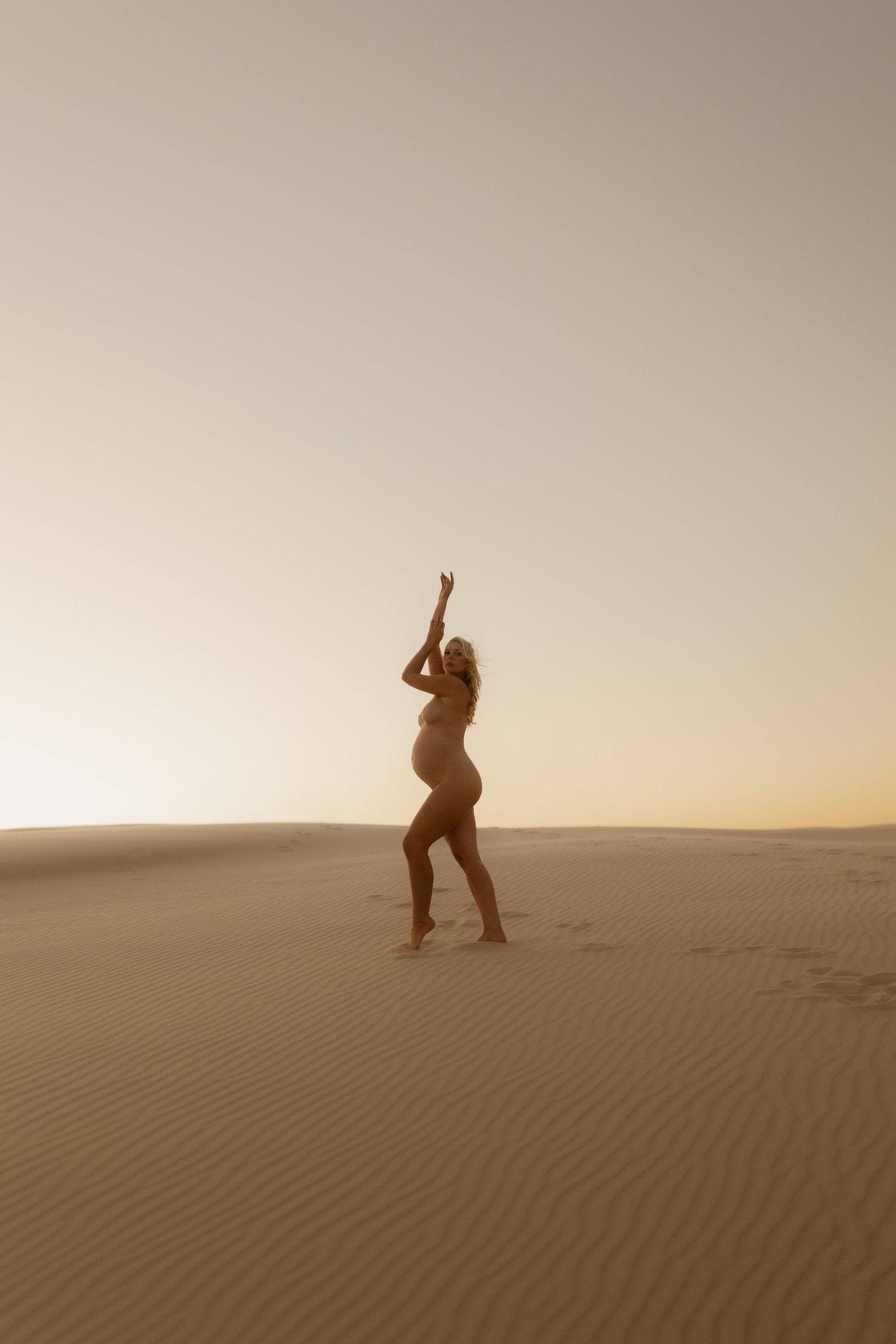 Pregnant woman in bikini posing with arms raised on golden desert sand dunes.