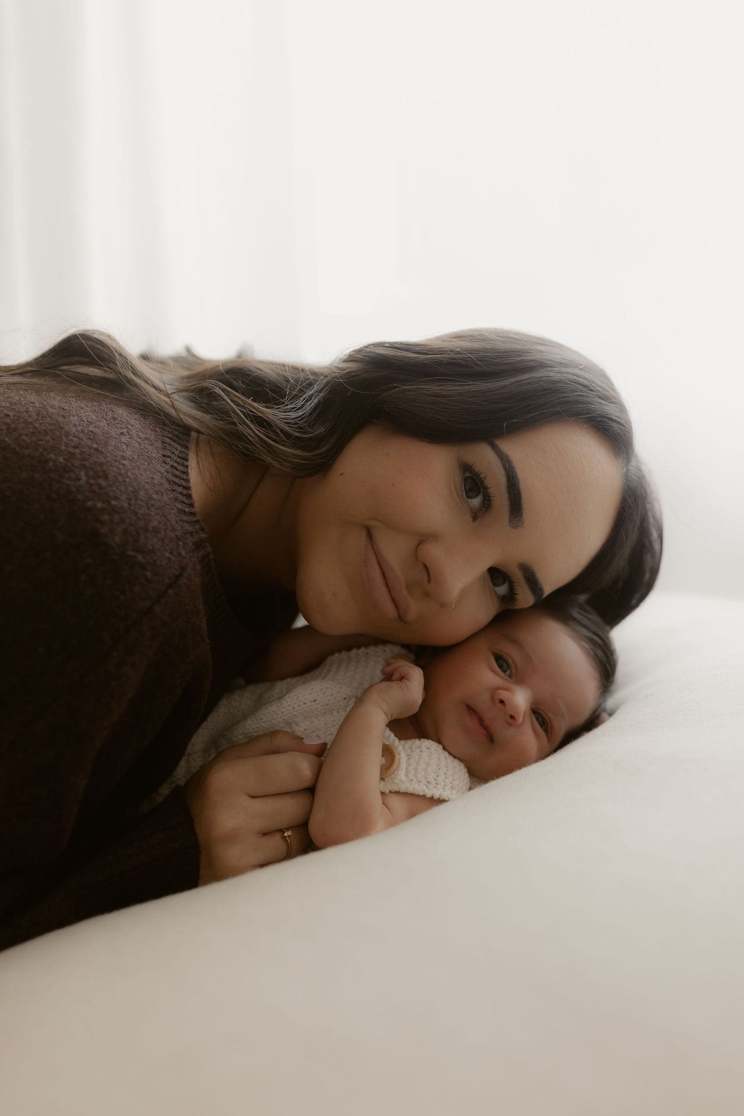 A mother tenderly rests her cheek against her newborn baby on a white surface in a soft, natural light setting.