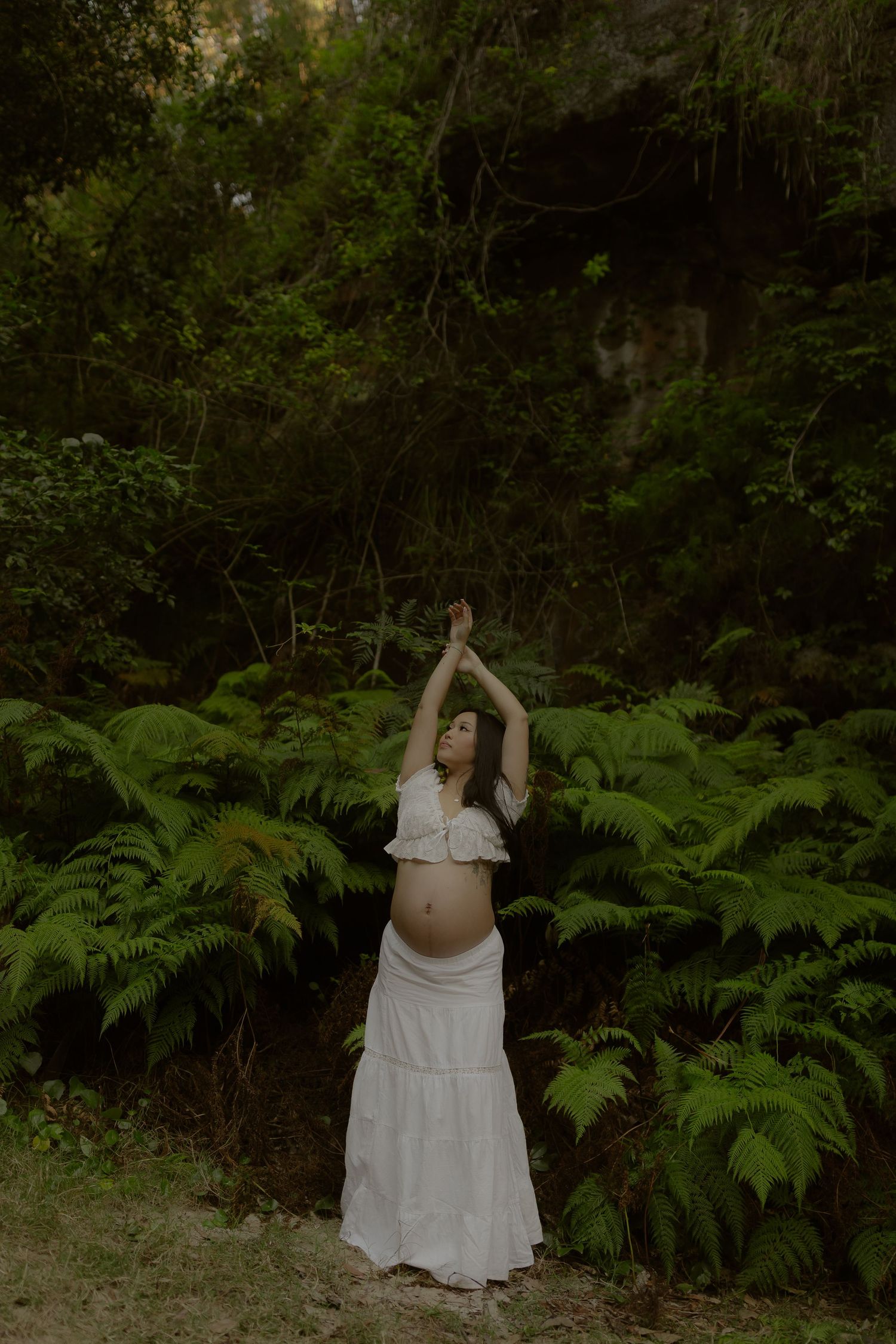 Pregnant woman in white outfit raising arms among green ferns in a lush forest setting.