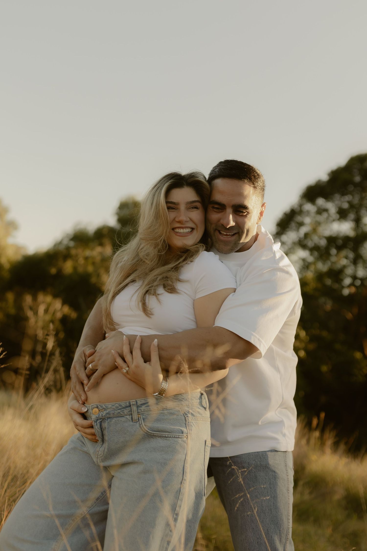 Smiling pregnant couple embracing in a golden sunlit field, wearing matching white tops and jeans.