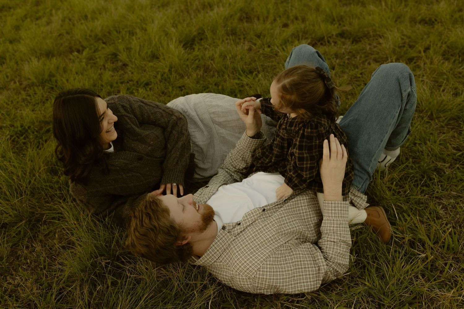 Family of three lying on green grass, laughing and bonding together outdoors in a candid moment.