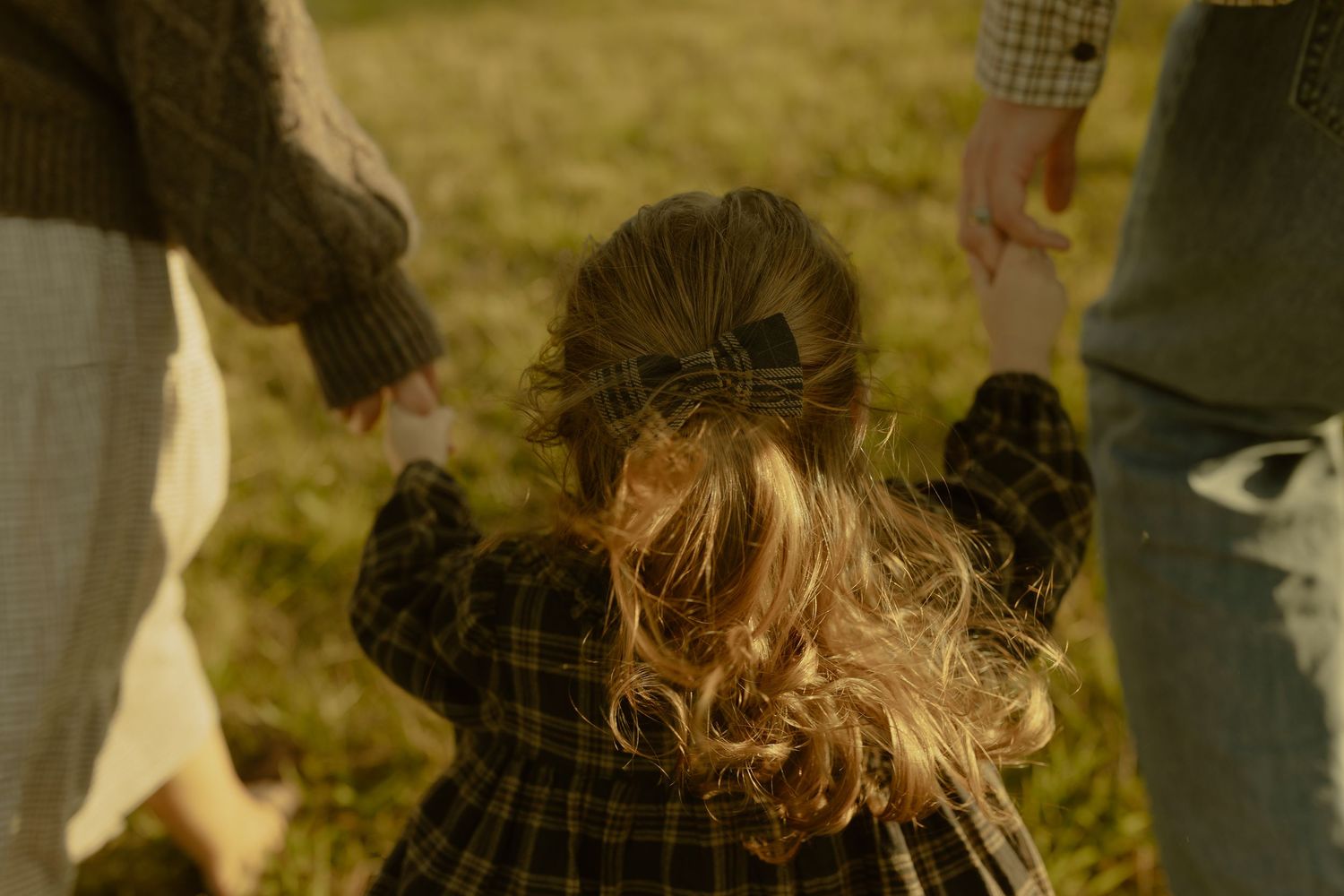 Young child with curly hair holds parents hands outdoors in golden light, wearing a plaid dress.