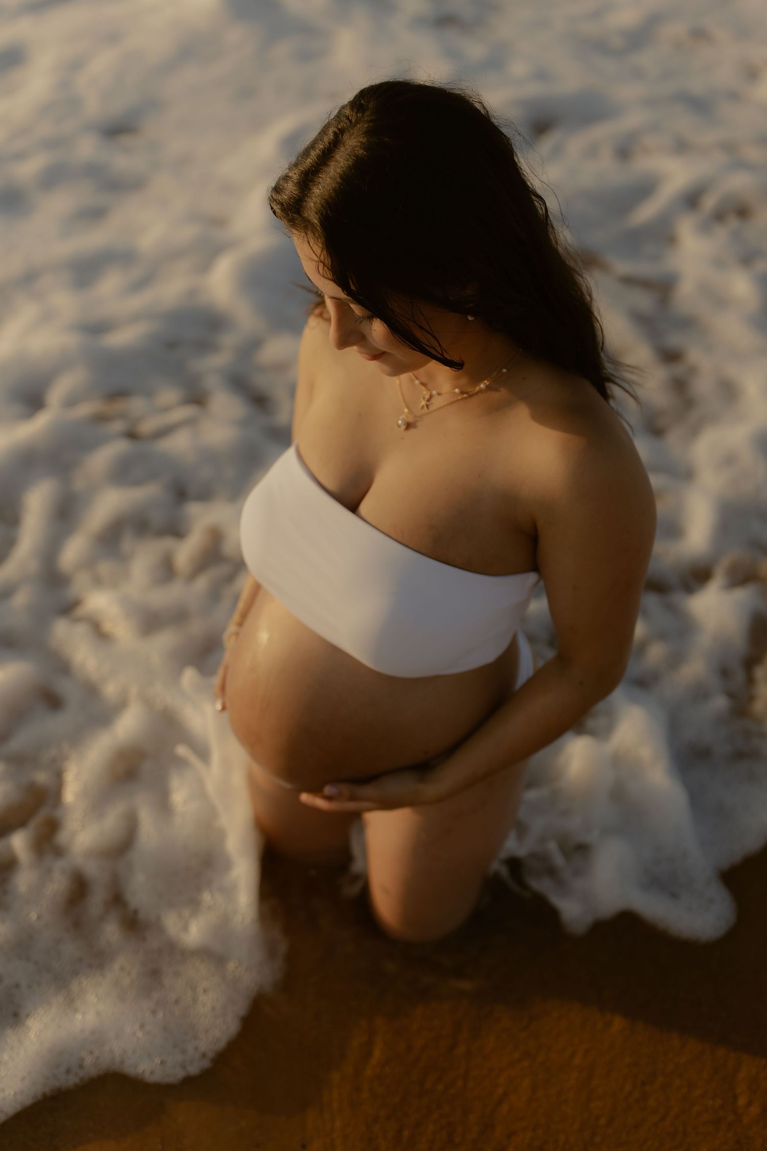 A serene maternity beach photoshoot captures an expectant mother sitting by the surf at sunset.