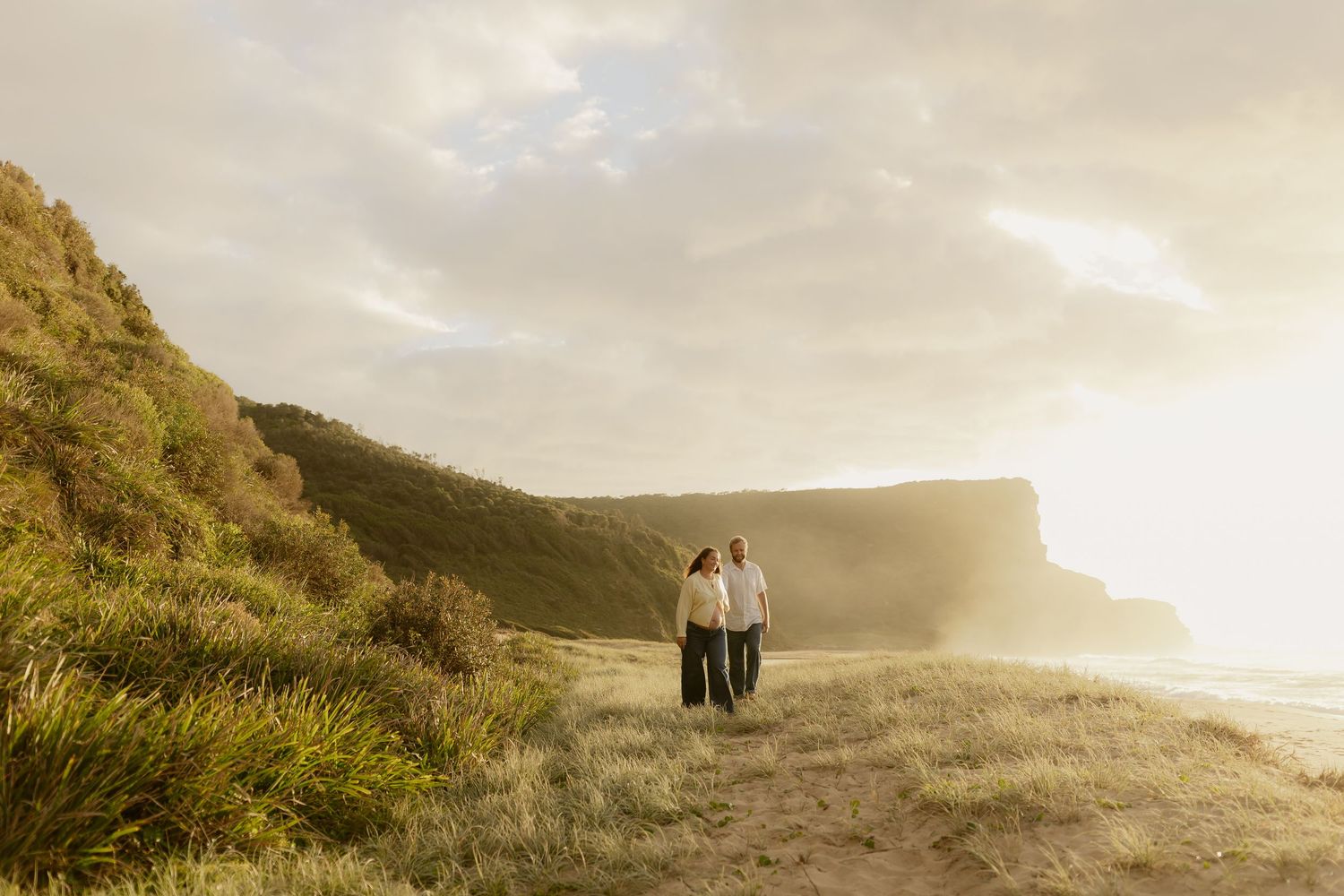 Two figures walk along a misty coastal cliff path at sunset with dramatic ocean views and golden light.