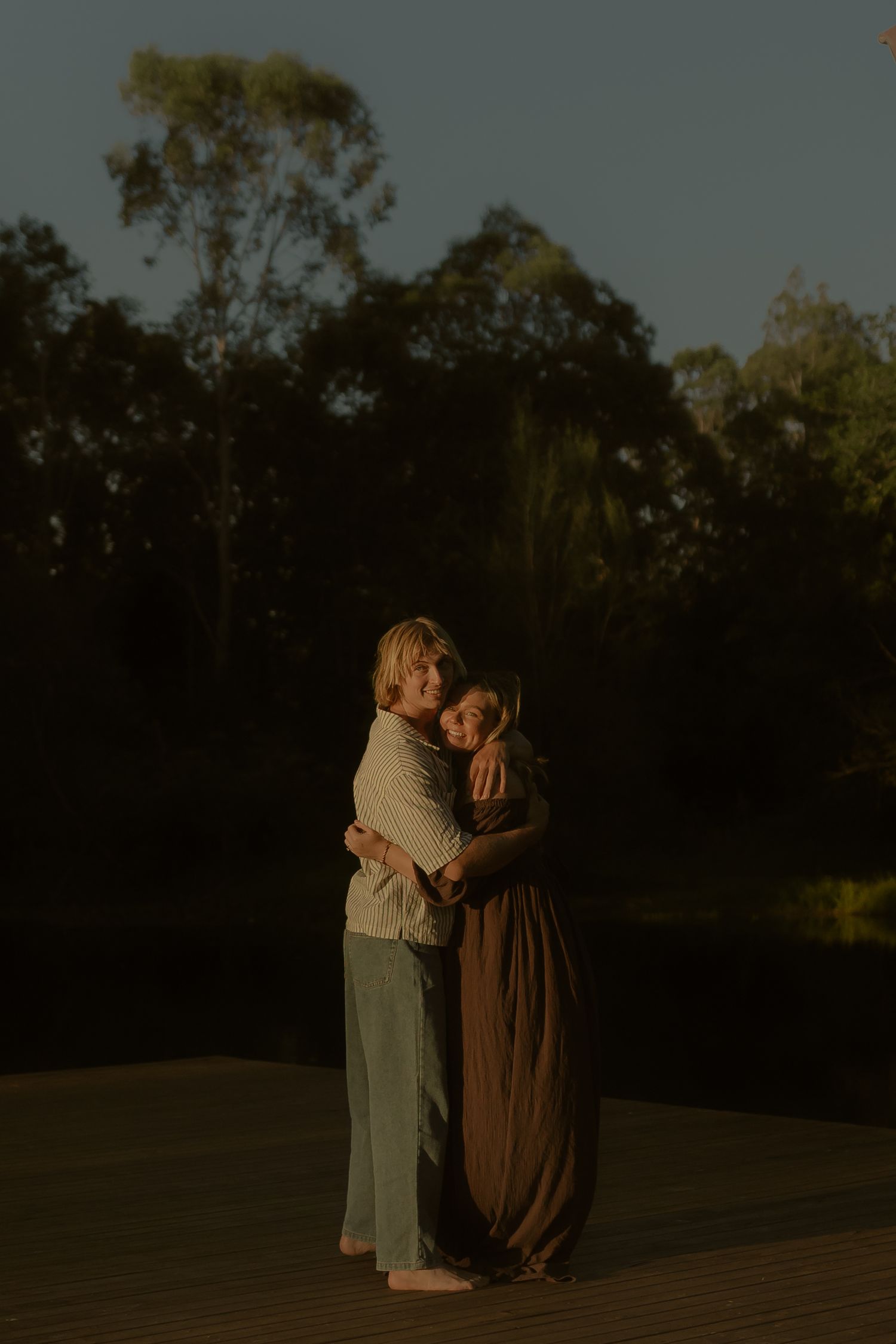 Two figures embrace on a wooden dock at sunset with trees silhouetted in the background.