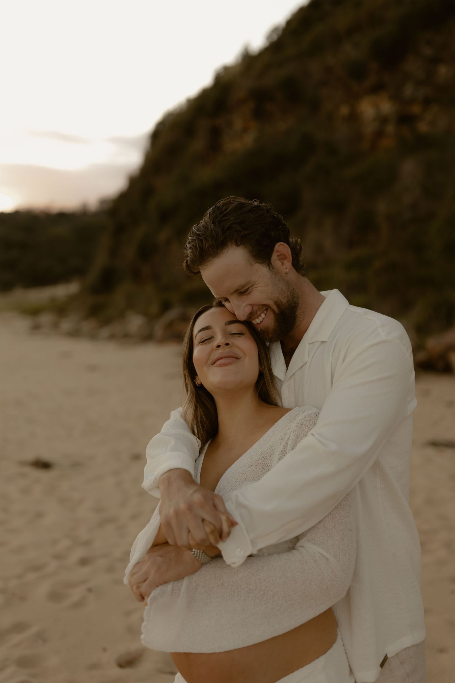 A couple embraces on a sandy beach at sunset with coastal cliffs in the background.