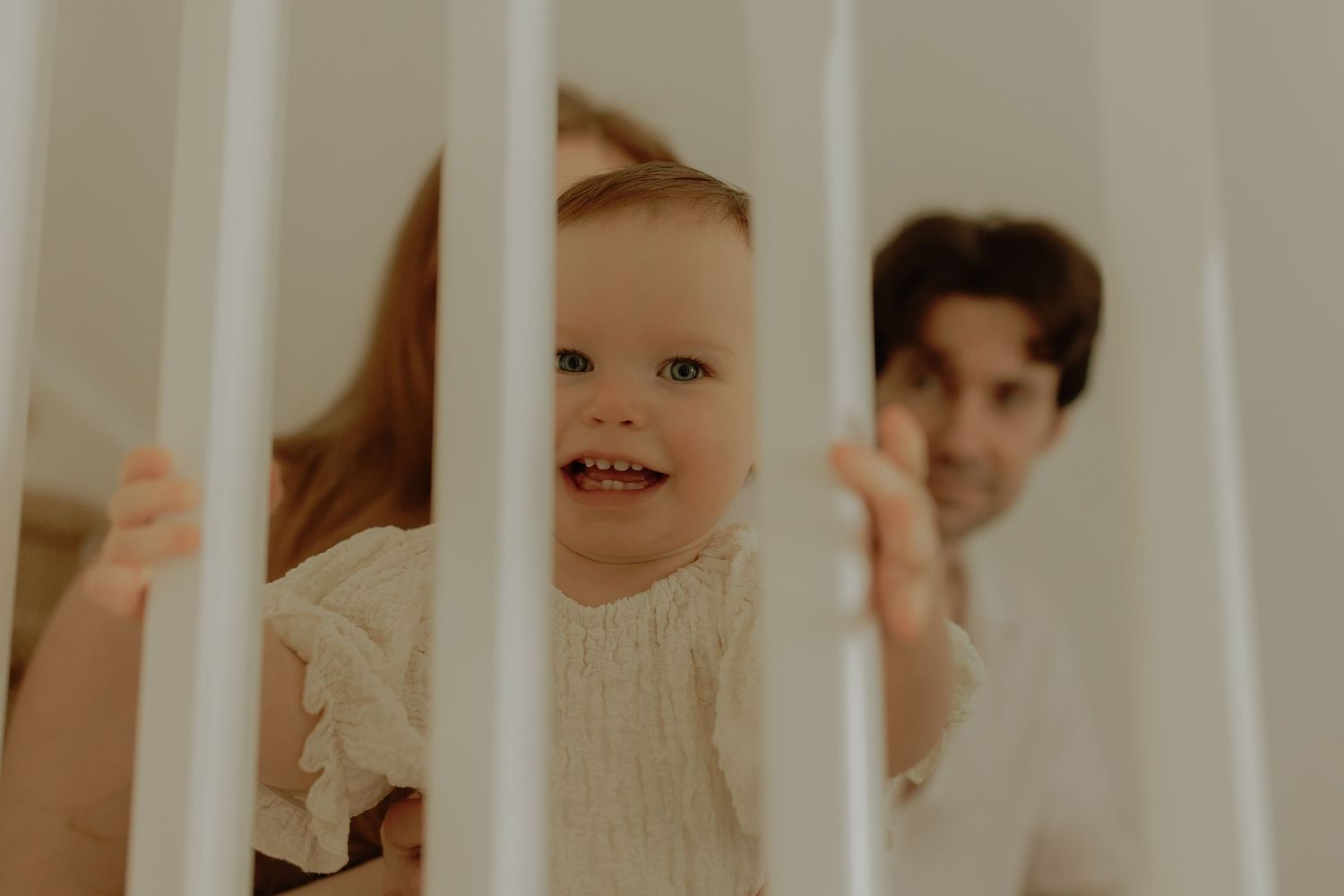 Happy toddler smiling through white crib rails with parents watching lovingly in soft natural light.