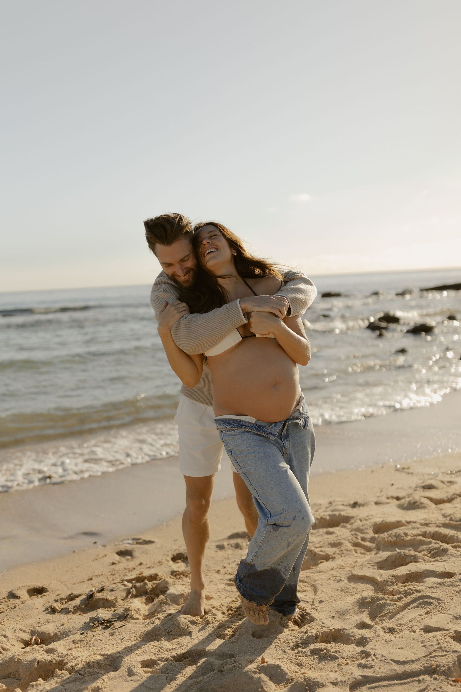 Couple plays and laughs together on sandy beach by ocean waves.