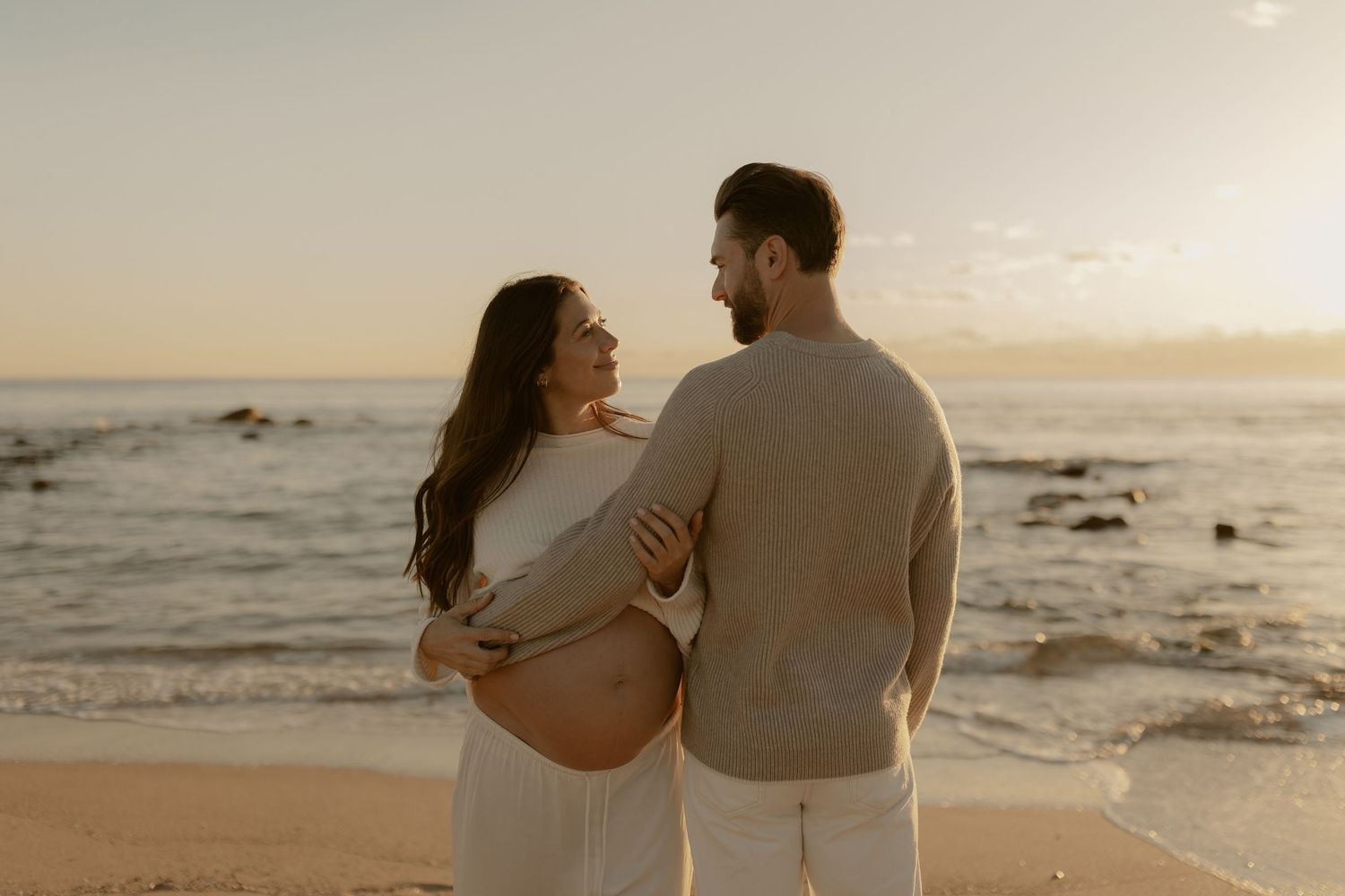 Romantic couple embracing on sandy beach during golden hour with ocean waves in background.