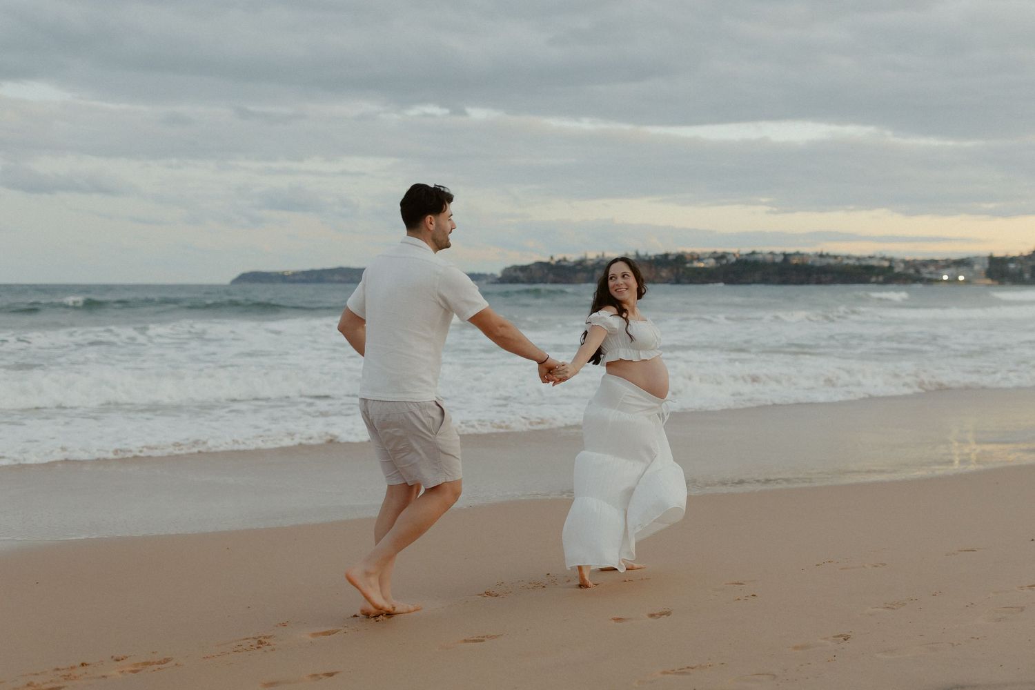 A couple in white outfits runs playfully along a sandy beach at sunset creating a romantic coastal scene.