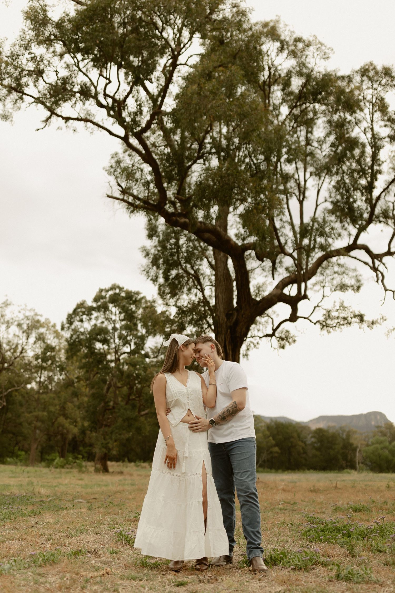 A couple embraces in a romantic outdoor setting with gum trees and mountains in the background.