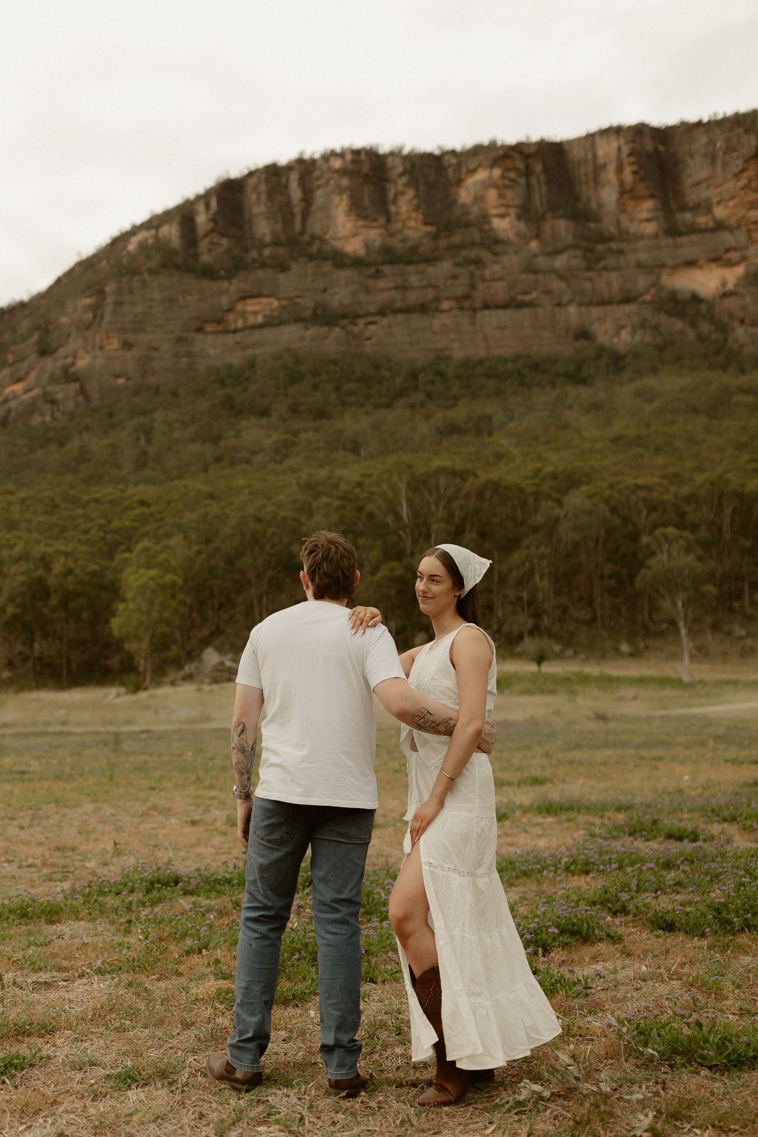 A couple stands together in a scenic landscape with dramatic cliffs and mountains in the background.