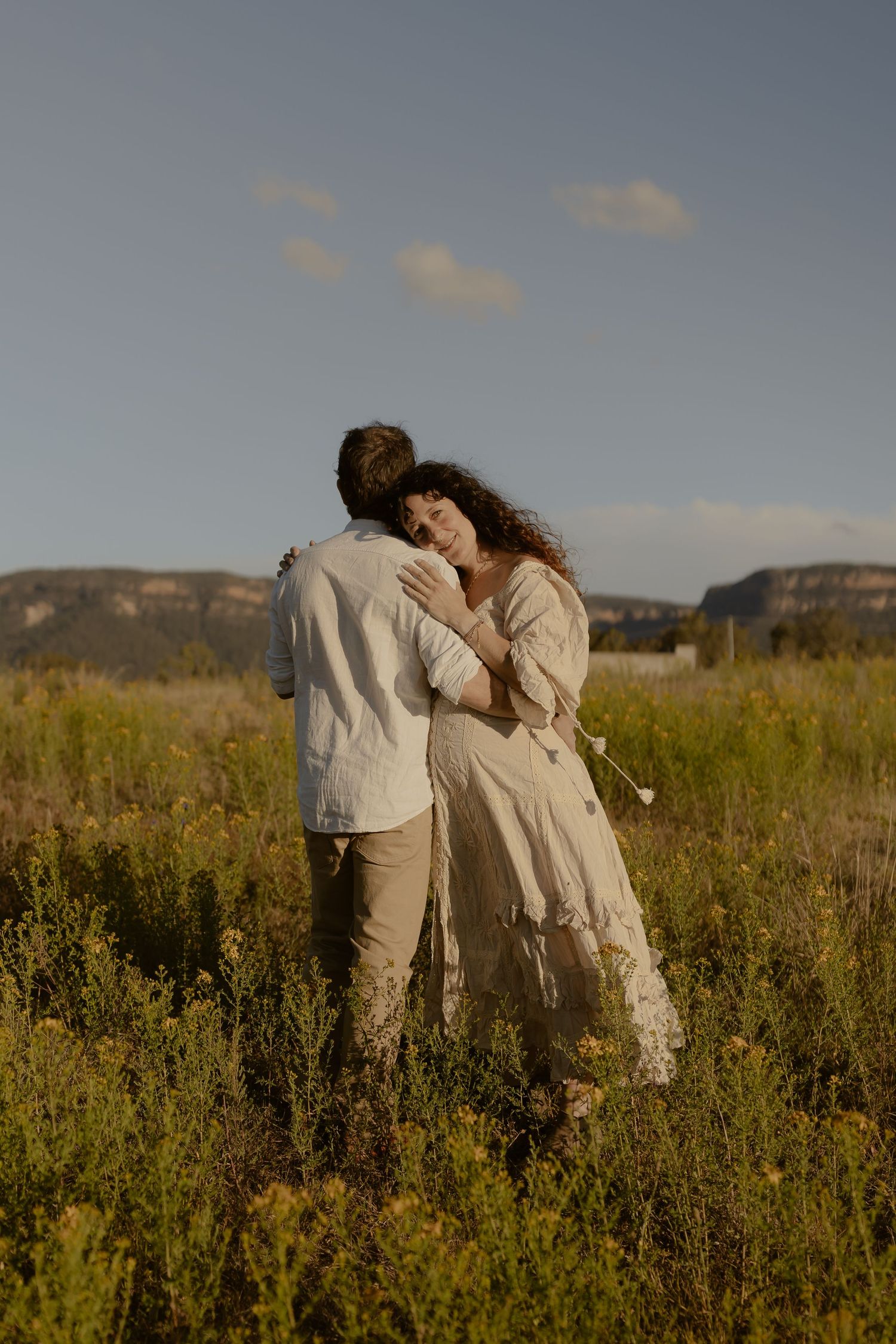 Two people share an intimate moment while standing among wildflowers in a mountain landscape.