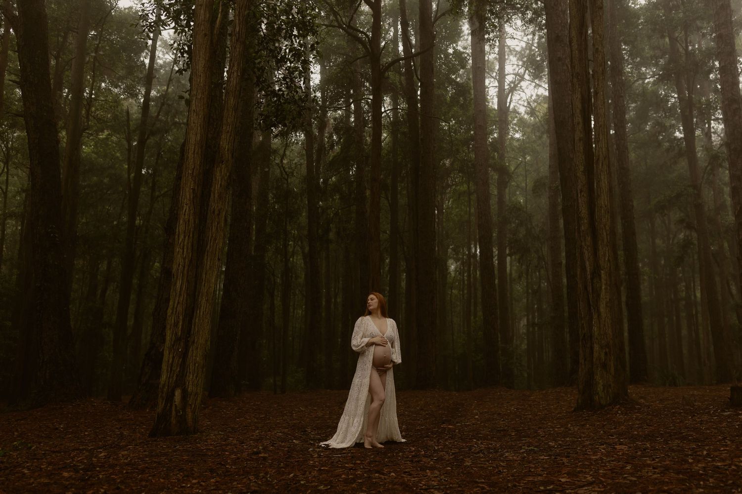 Woman in white gown standing among tall trees in a misty, dark forest with red leaf-covered ground.