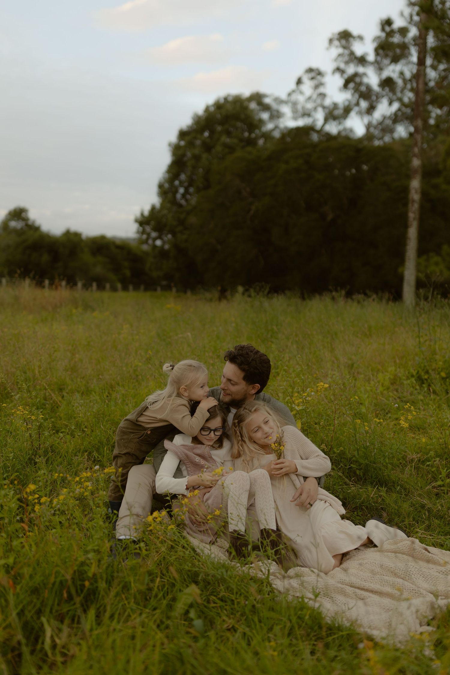 A family of four sits together in a wildflower meadow at dusk, sharing a tender moment.