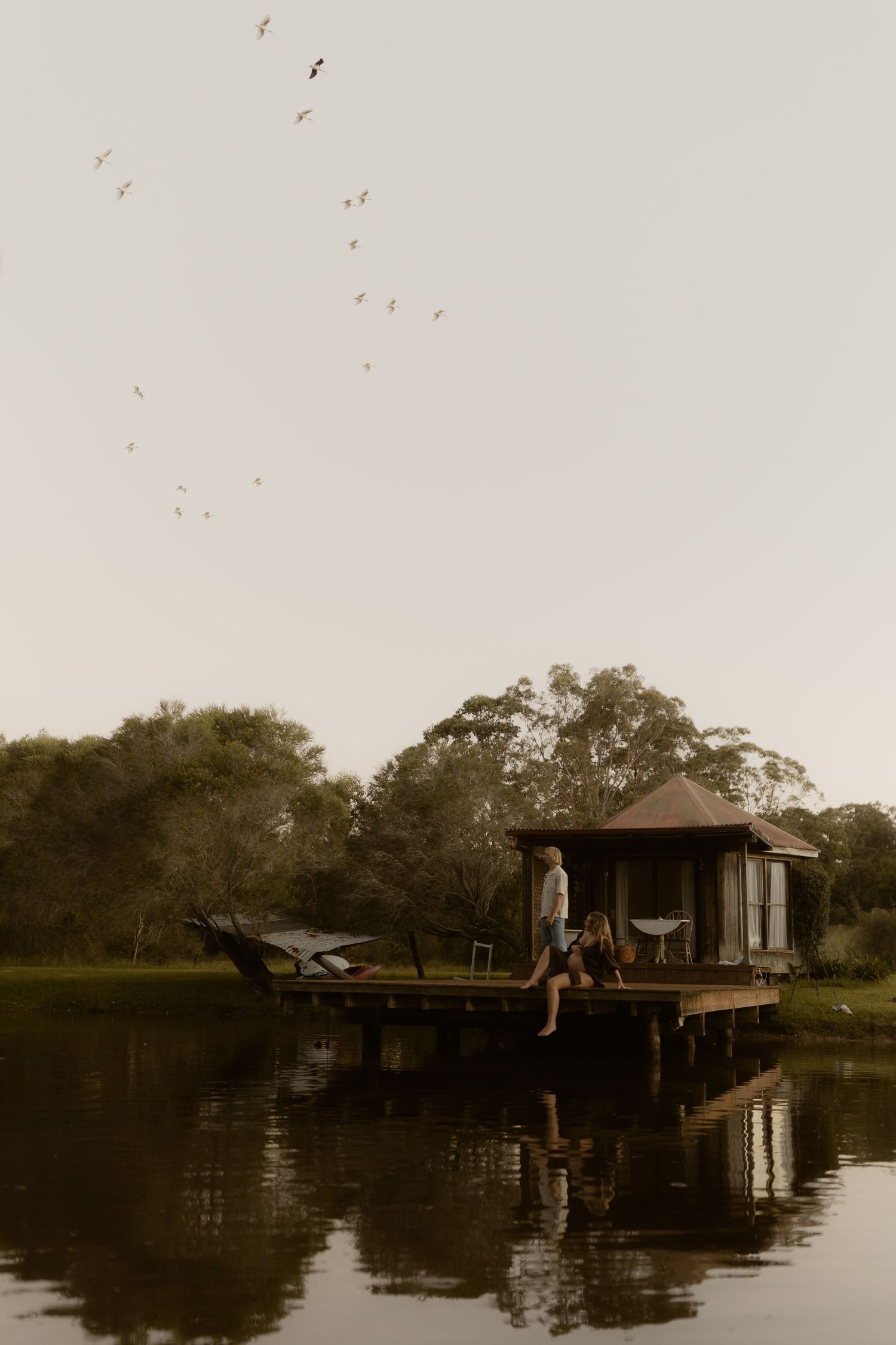 Person sitting on a floating dock with a small cabin over a tranquil lake at dusk, birds flying above.