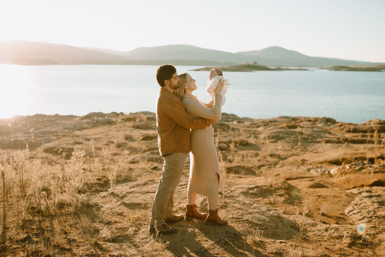 A couple embraces on a rocky cliff overlooking a serene lake and mountain landscape during golden hour.