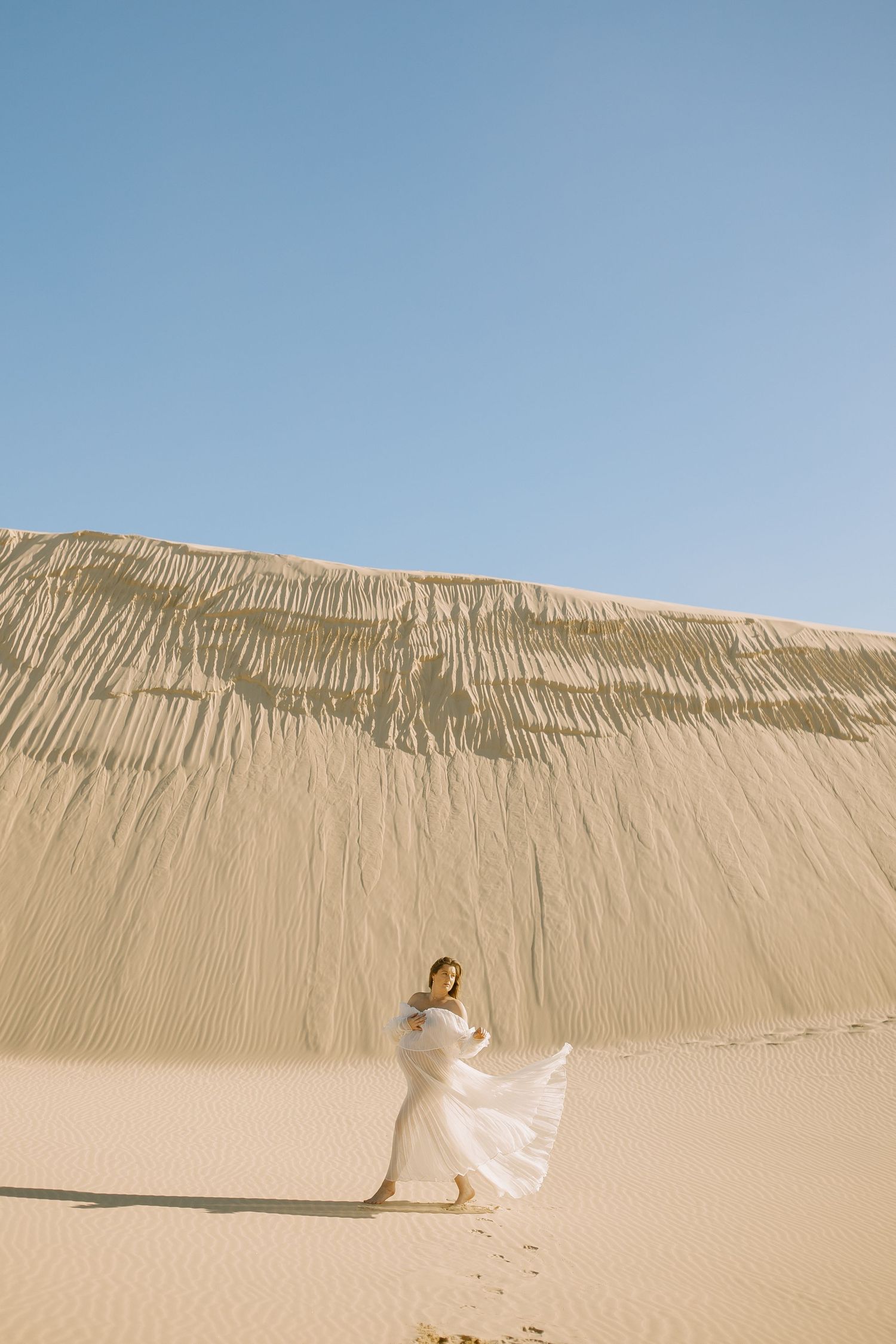 A figure in flowing white fabric performs a graceful dance sequence across a vast desert sand dune landscape.
