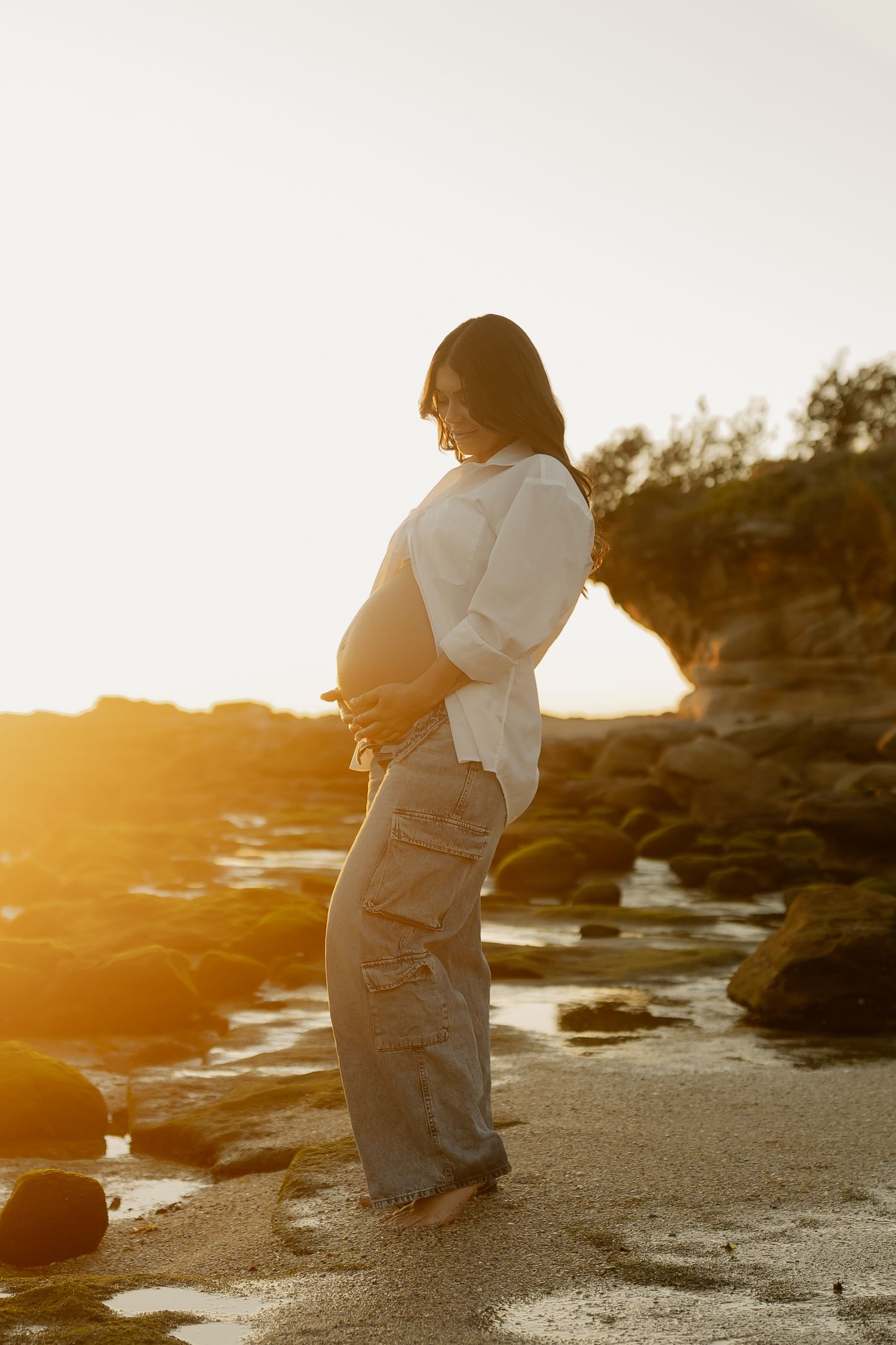 Pregnant woman cradling her baby bump on a rocky beach at golden sunset, wearing white shirt and grey cargo pants.