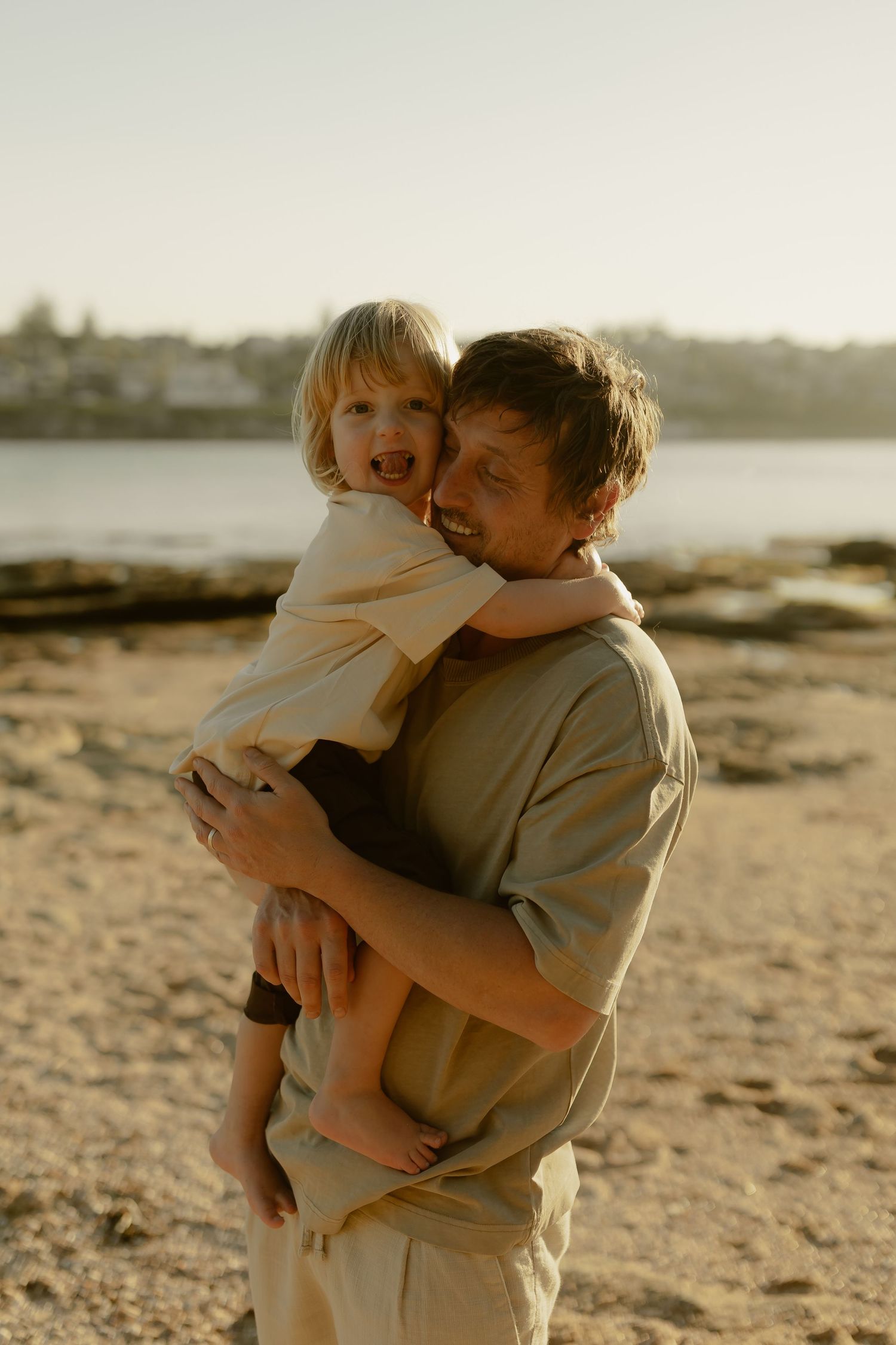 A smiling father holds his laughing toddler on a sandy beach during warm golden hour light.
