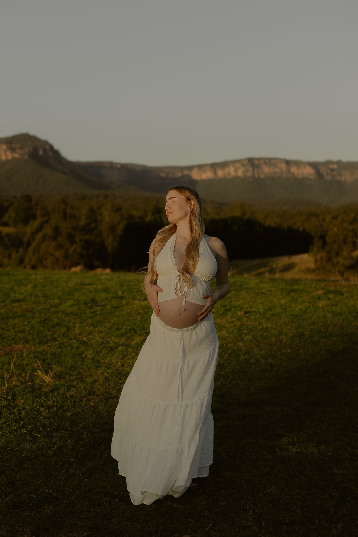 Pregnant woman in white halter maxi dress cradling baby bump at golden hour with mountain backdrop.