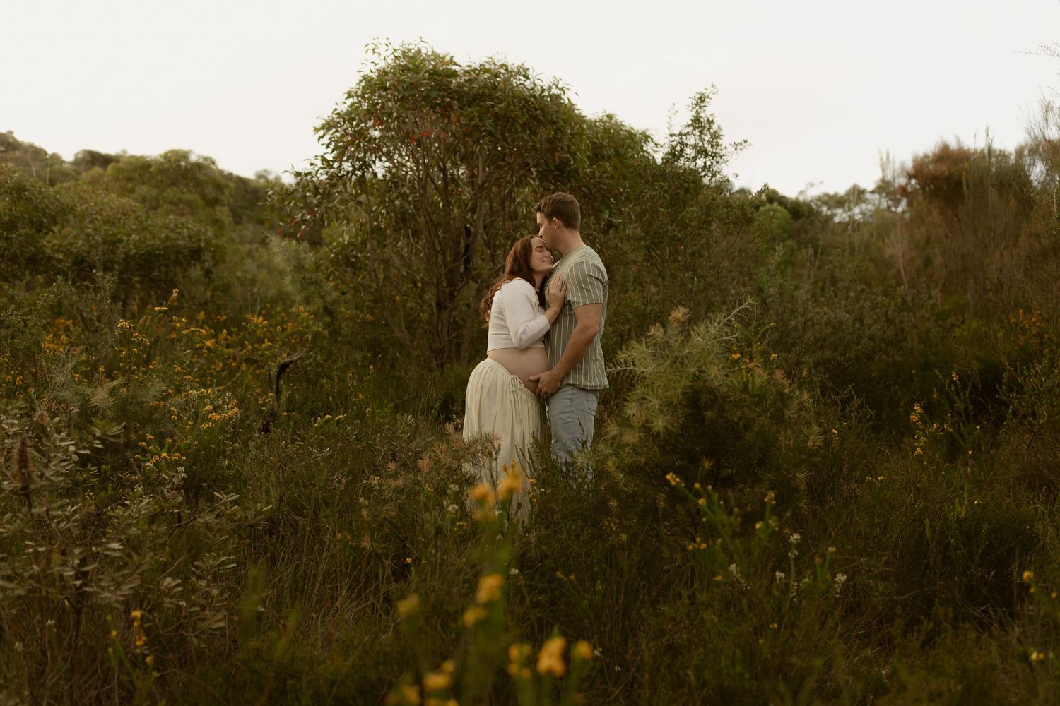 A couple embraces romantically among lush wildflowers and greenery in a serene outdoor setting.