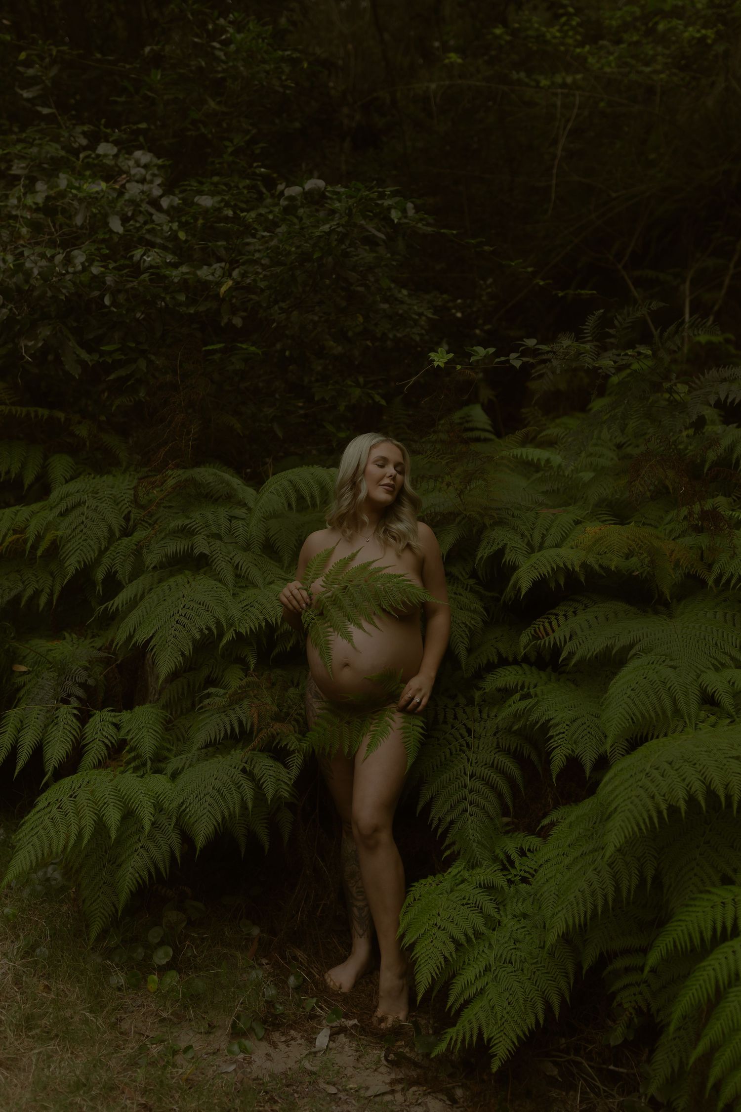 Pregnant woman posing nude among green ferns in a dark forest, smiling softly.