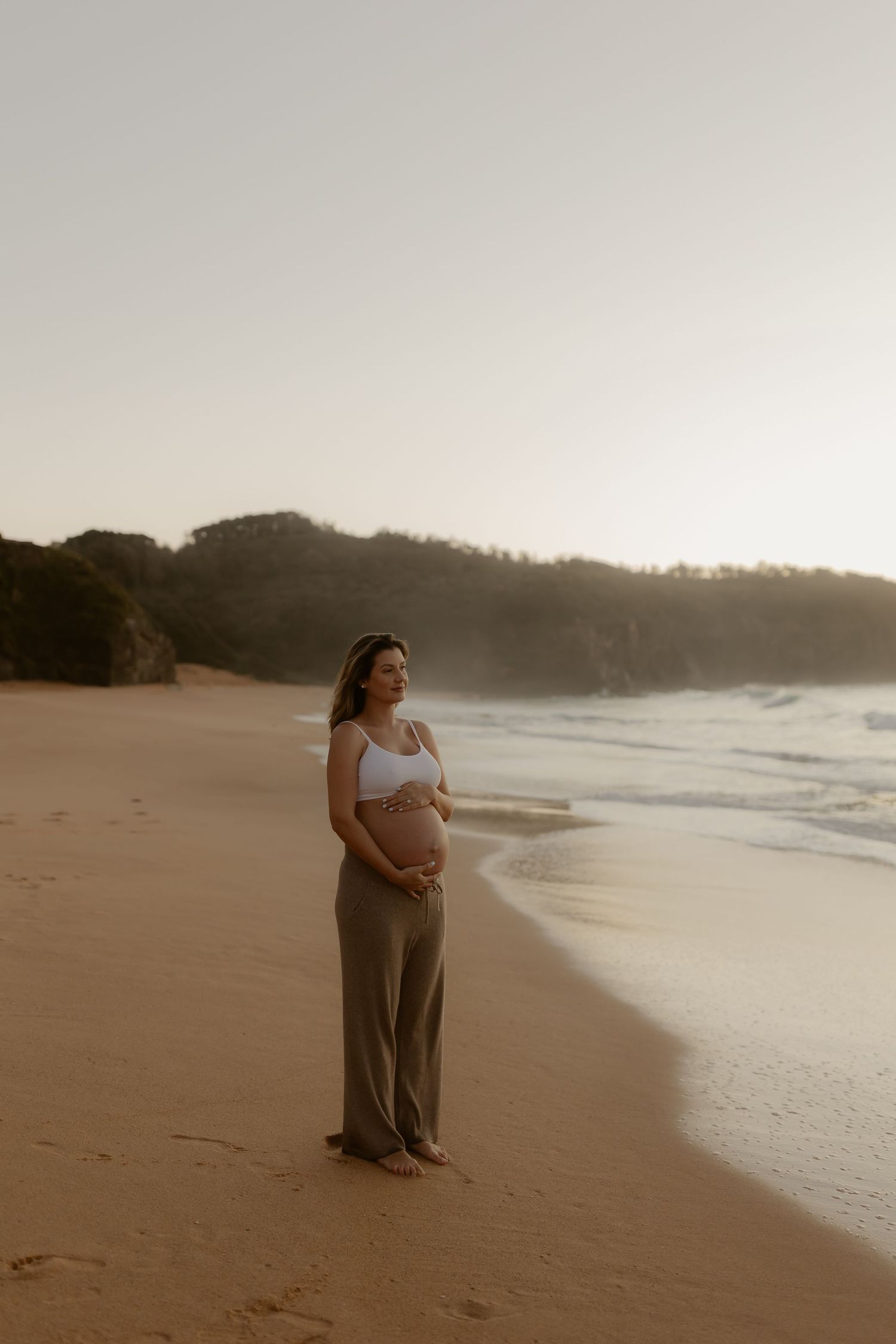 Pregnant woman in white crop top and olive pants cradling baby bump on sandy beach at sunset.