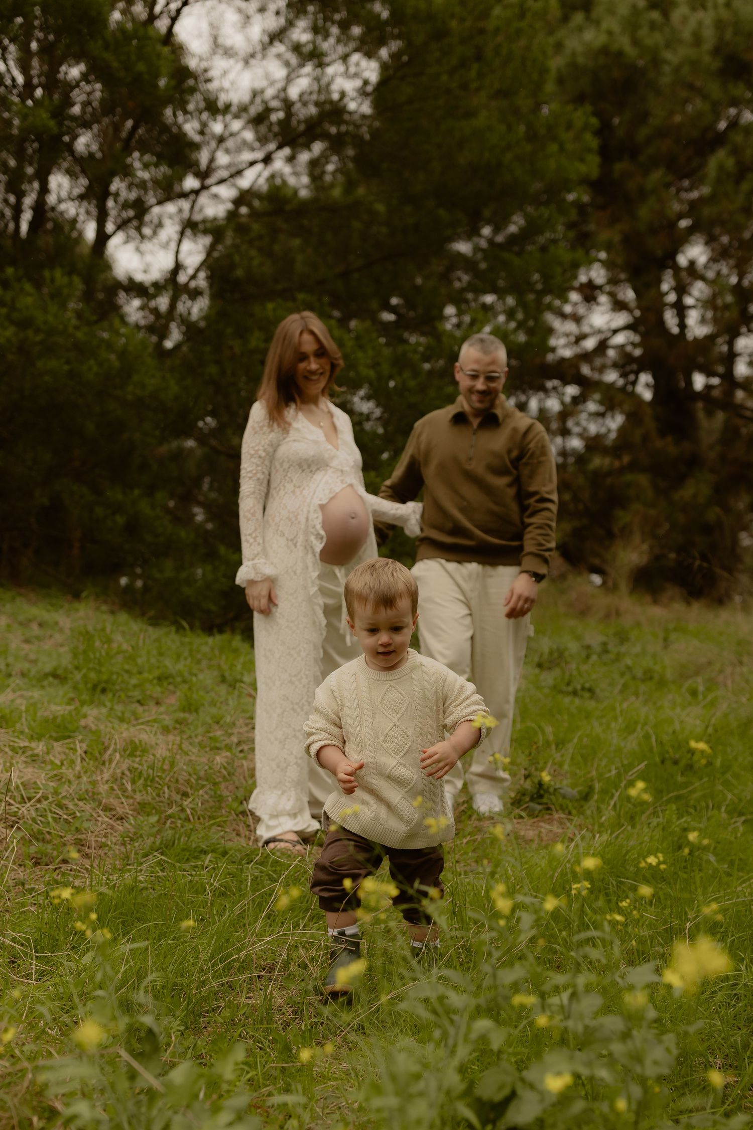 Family maternity photo with pregnant woman, partner, and toddler in green meadow.