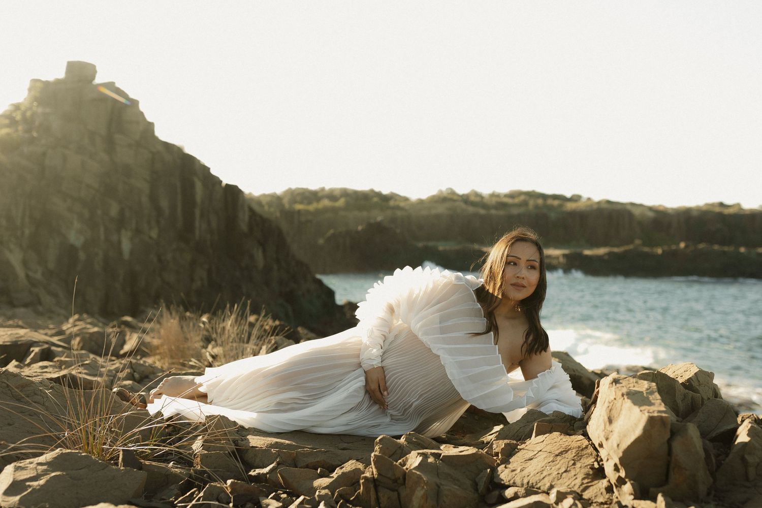 A woman in white dress poses on coastal rocks with ocean waves in the background.