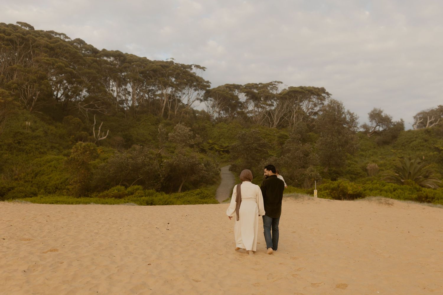 Couple walking hand in hand on sandy beach toward lush green forest under overcast sky.