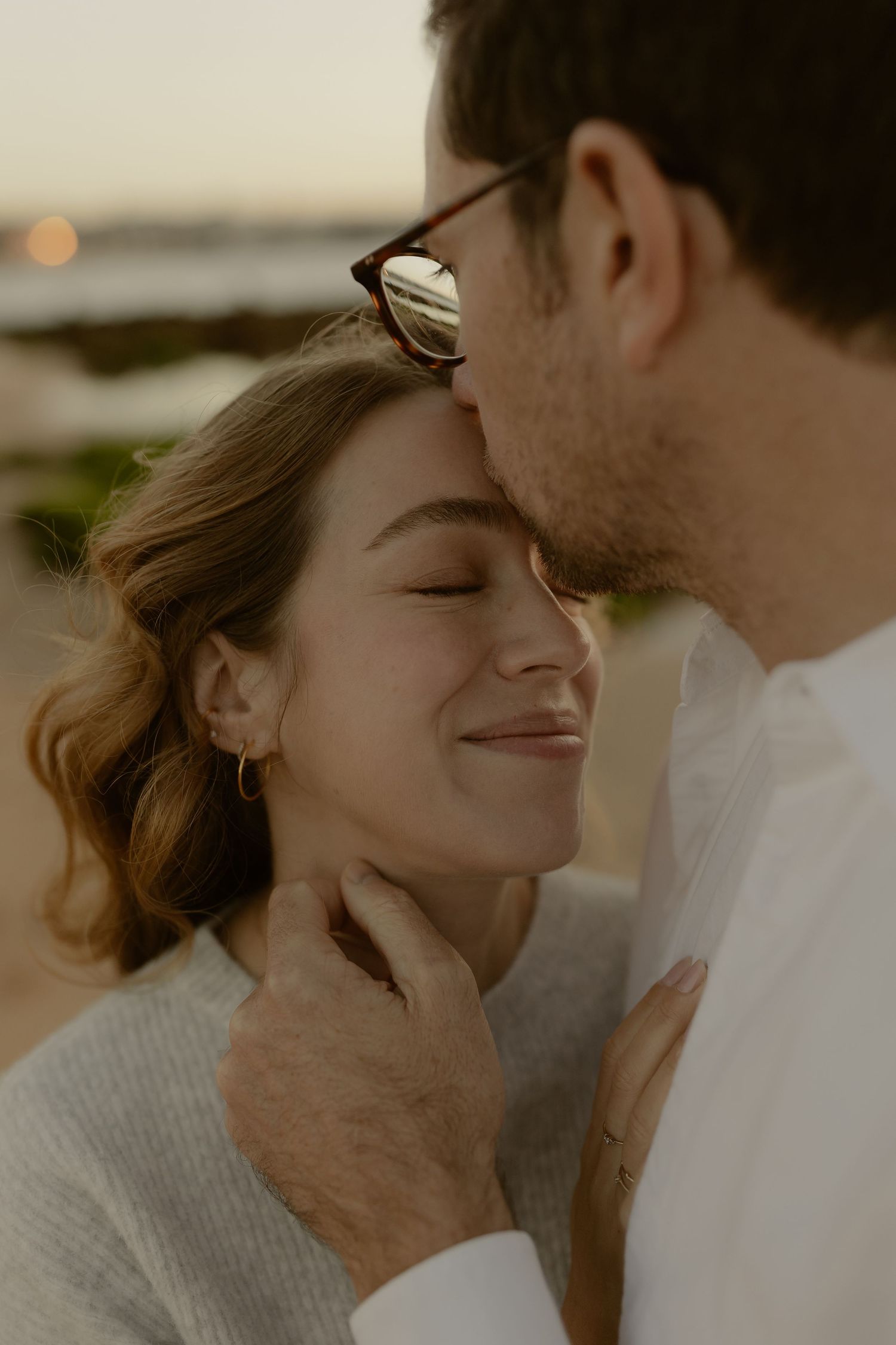 Tender moment as man kisses pregnant woman's forehead on beach at sunset, both eyes closed.